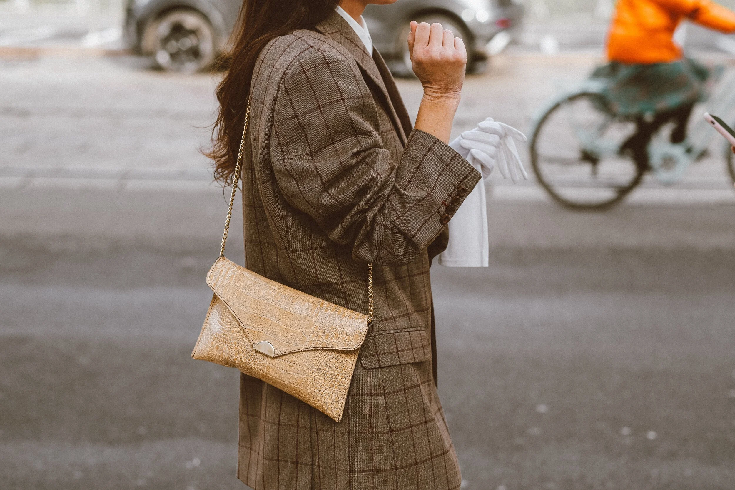 A woman wearing a brown plaid blazer with a beige textured clutch bag over her shoulder, holding a white cloth and talking to someone on the street.