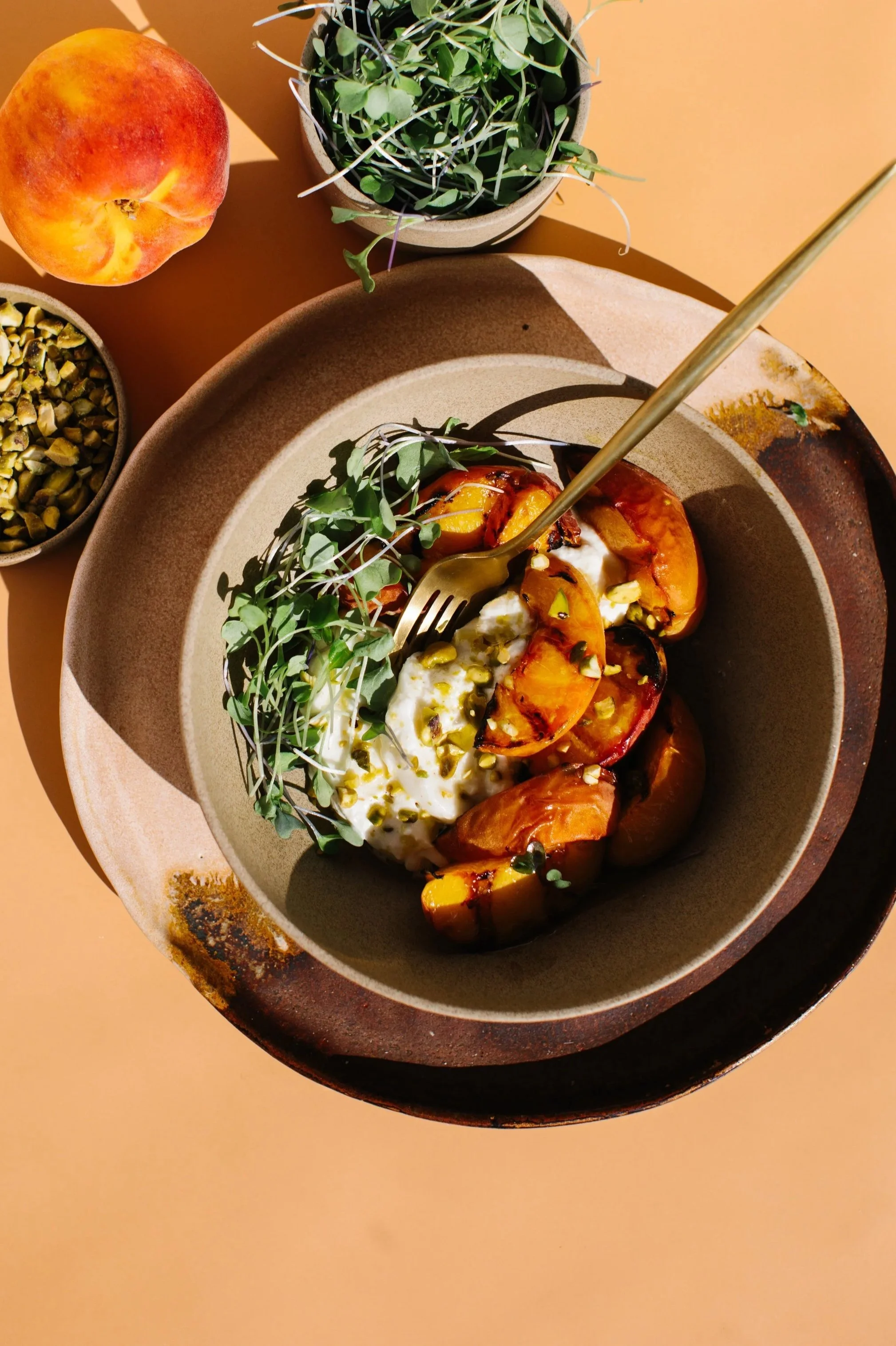 A bowl of grilled peaches topped with yogurt, chopped pistachios, and microgreens on a ceramic plate, with a peach, a bowl of pistachios, and a potted plant on an orange table.