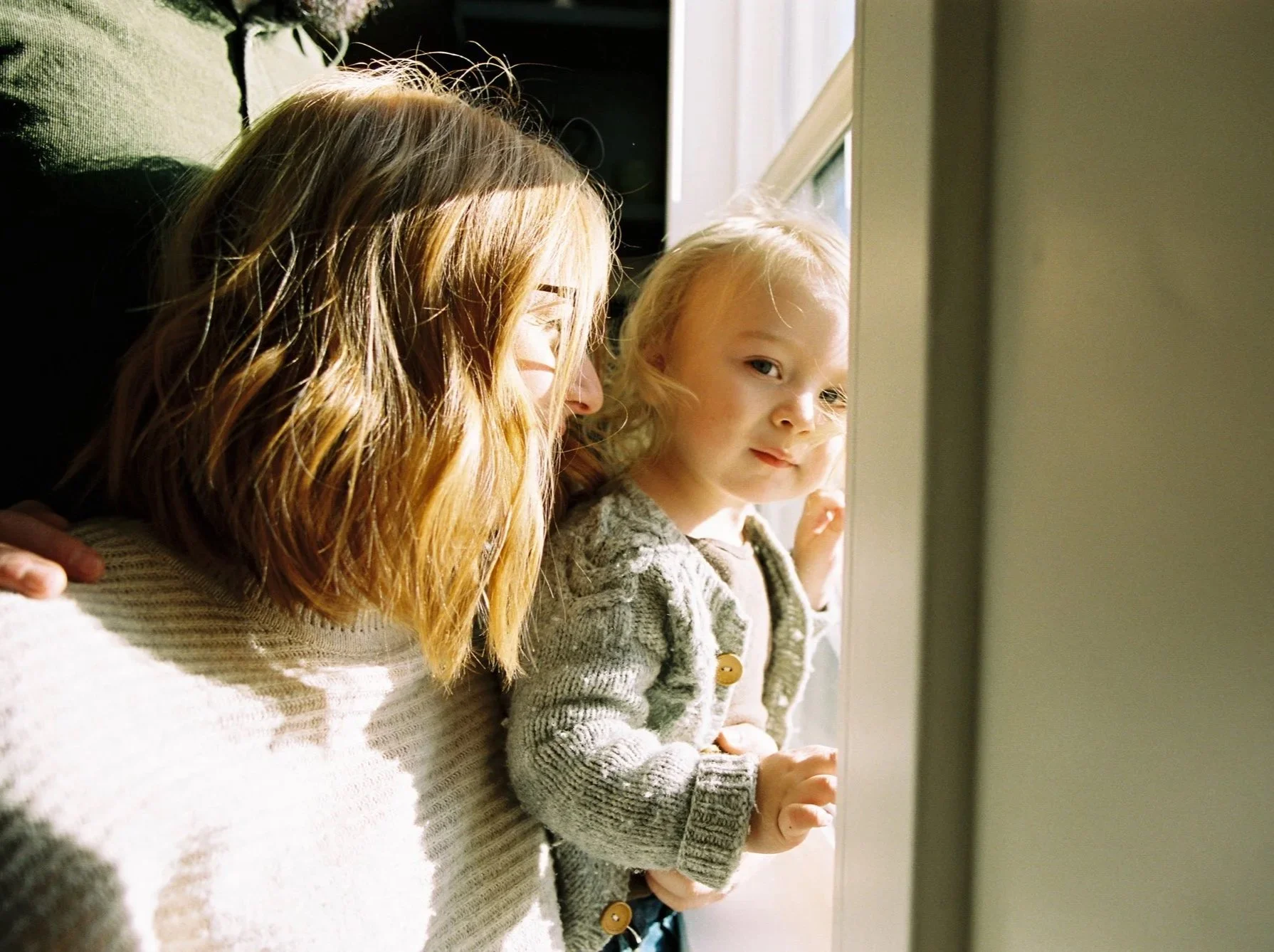 A woman with reddish hair and a young girl with blonde hair looking out a window, with natural sunlight illuminating their faces.
