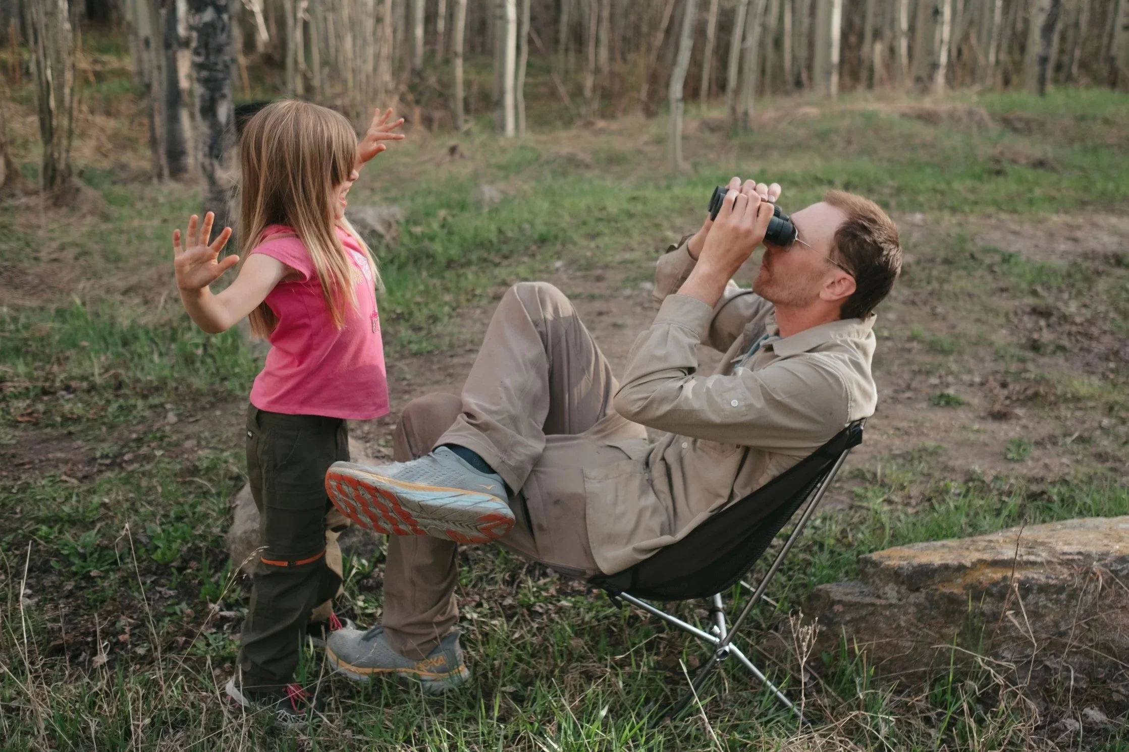 A man sitting in a chair using binoculars to look into the distance as a young girl in a pink shirt and dark pants stands nearby, gesturing with her hands in a forest setting.
