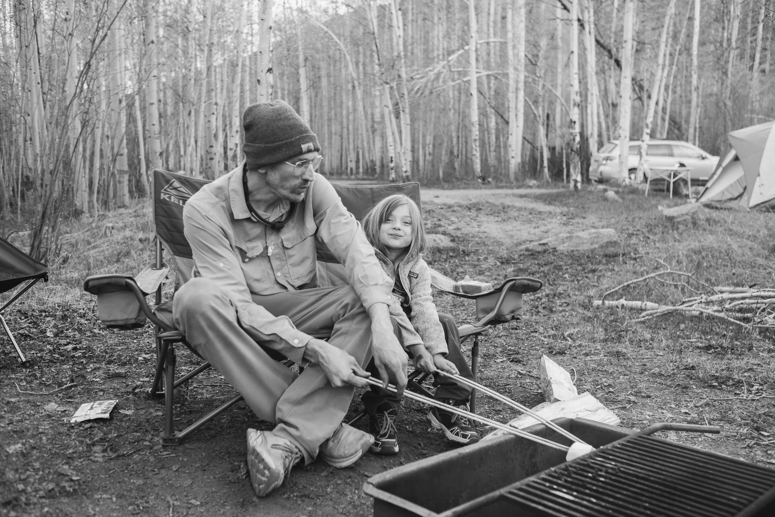 A man and a young girl sit on a camping chair outdoors in a wooded area. The man is preparing to cook on a grill, and the girl is smiling. There are tents, a car, and trees in the background.