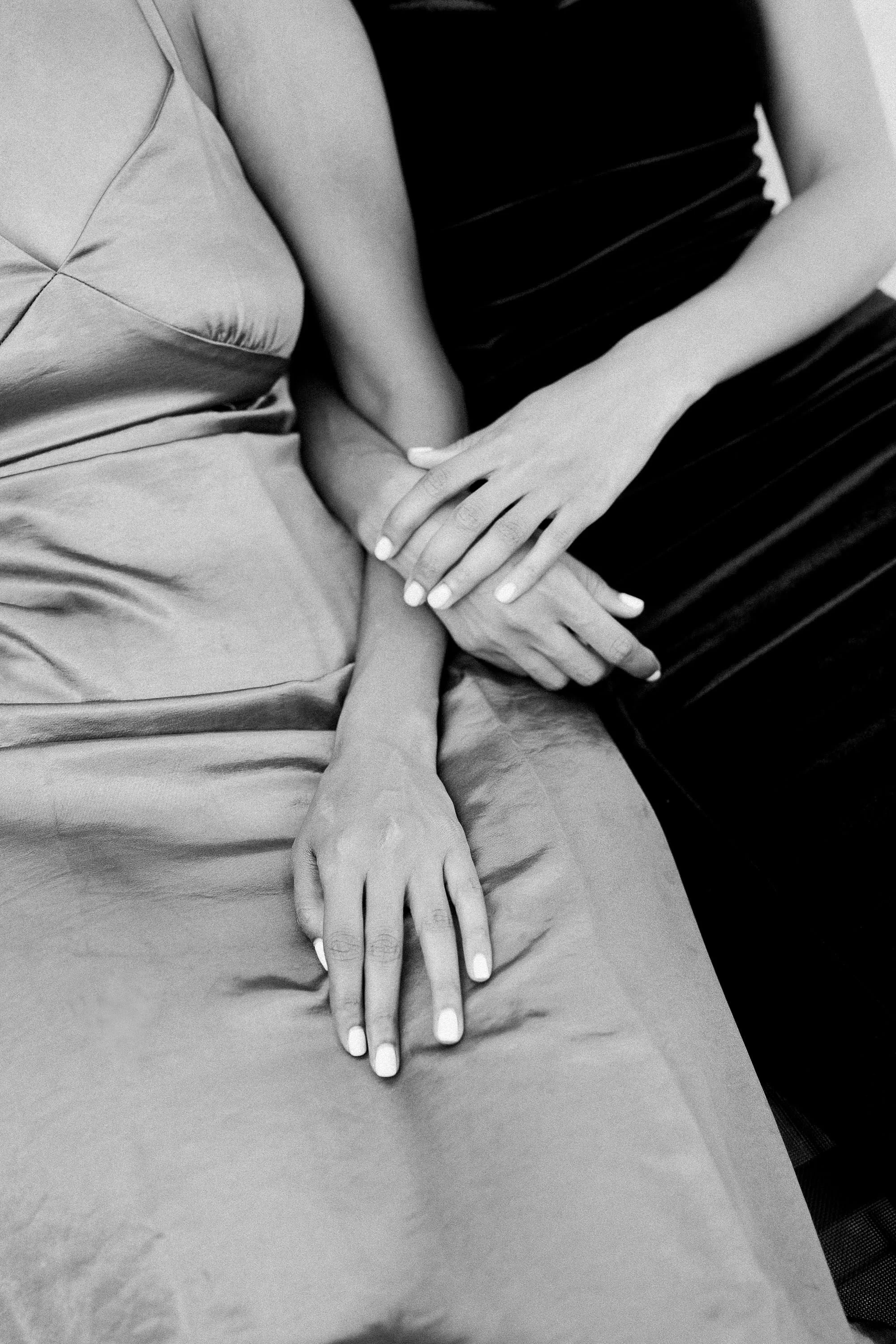 Close-up of two women holding hands, dressed in formal attire; one in a satin dress and the other in a dark dress.