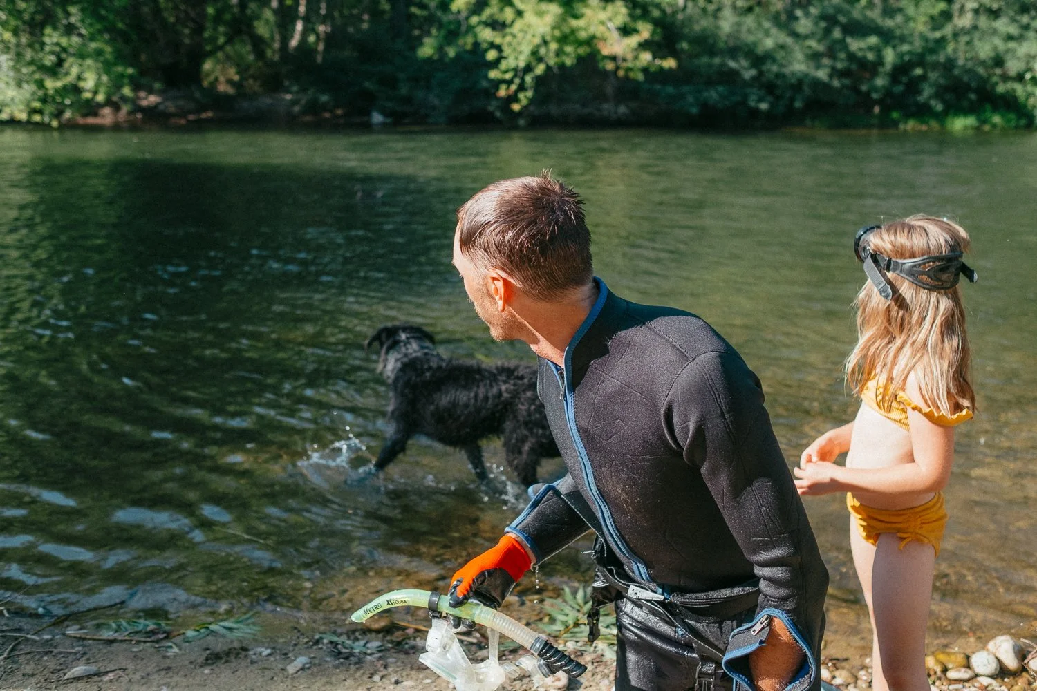 A man and a young girl on a riverbank in Boise, Idaho., with an inflatable snorkel mask, near a black dog swimming in the river, surrounded by trees.
