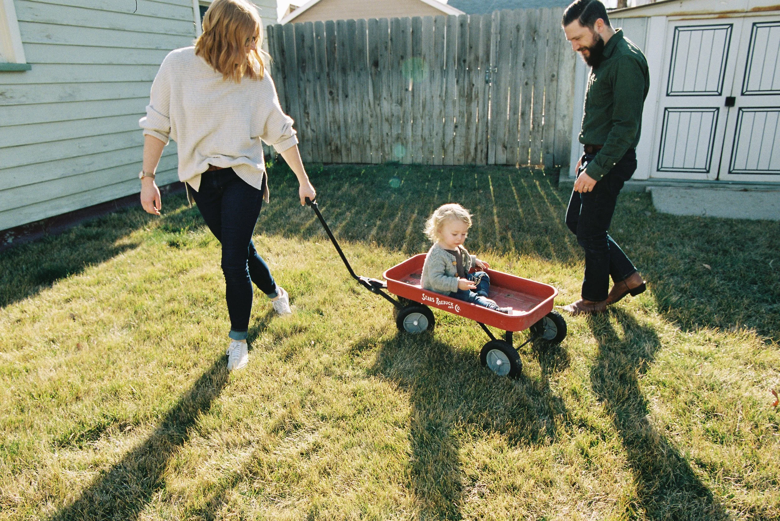 A family of three playing in a backyard in Boise, Idaho, with a woman pulling a red wagon carrying a young girl while a man stands nearby.