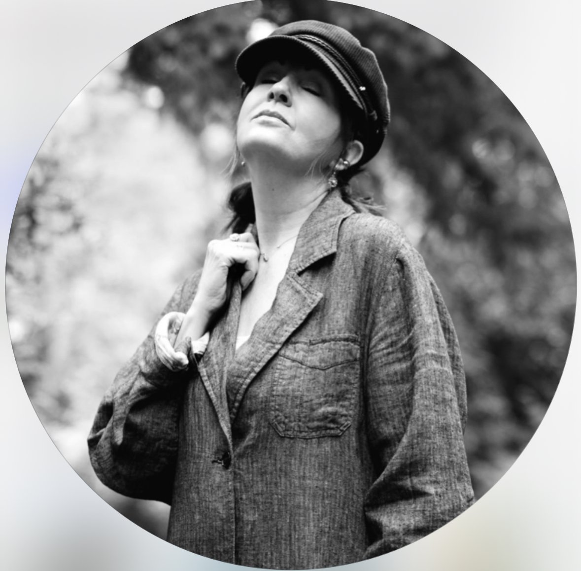 Black and white photo of a woman outdoors wearing a cap and a casual jacket, looking confidently upward with her hand on her shoulder.