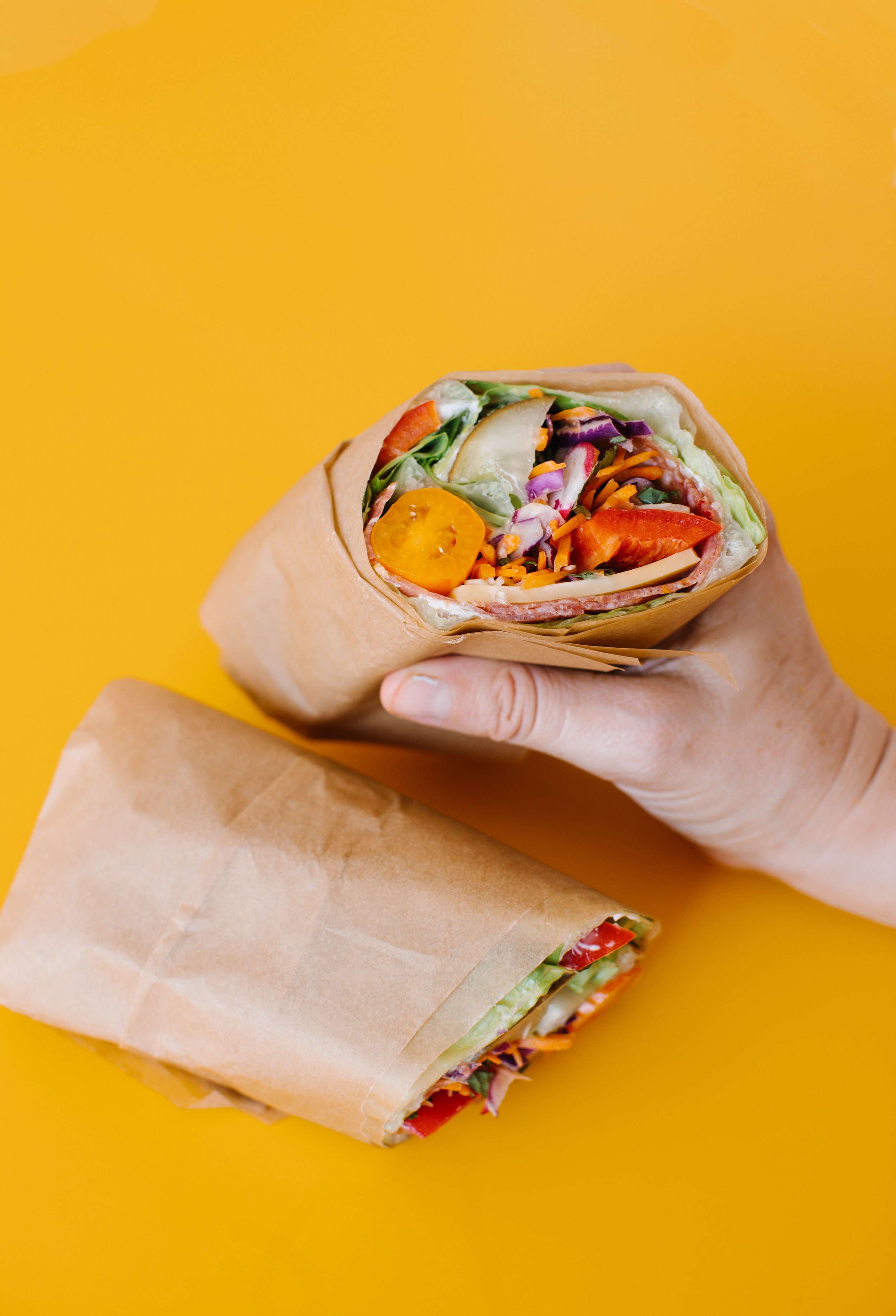 A hand holding a vegetable and meat wrap on a yellow surface, with another wrap lying below.