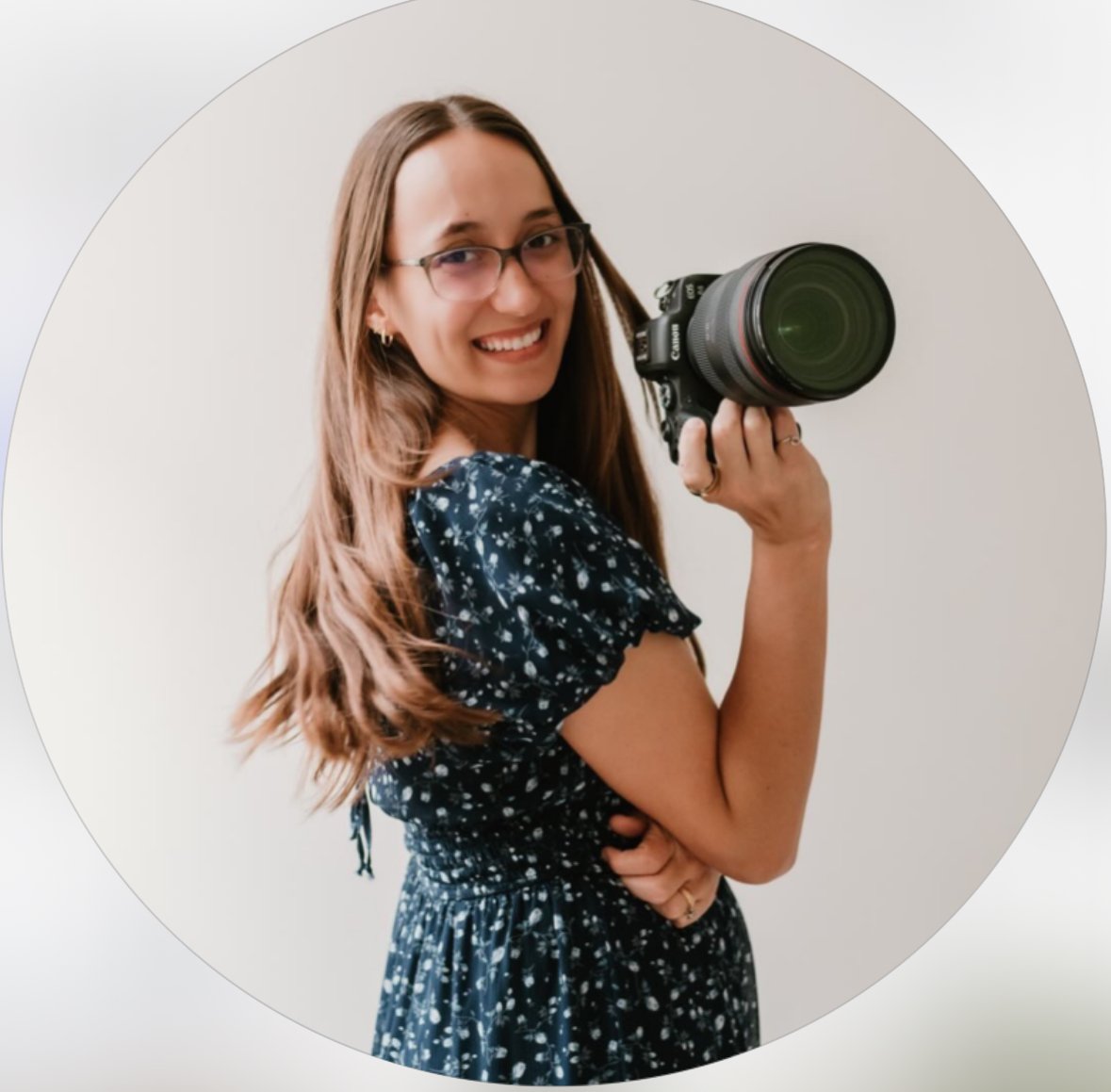 A smiling woman with glasses holding a professional camera, wearing a dark floral dress, standing against a plain light background.
