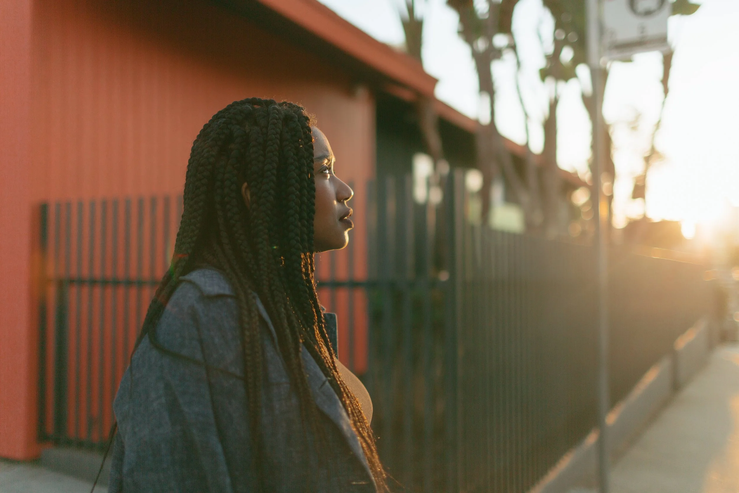 A woman with braided hair standing near a fence, looking towards the sunlight during sunset.