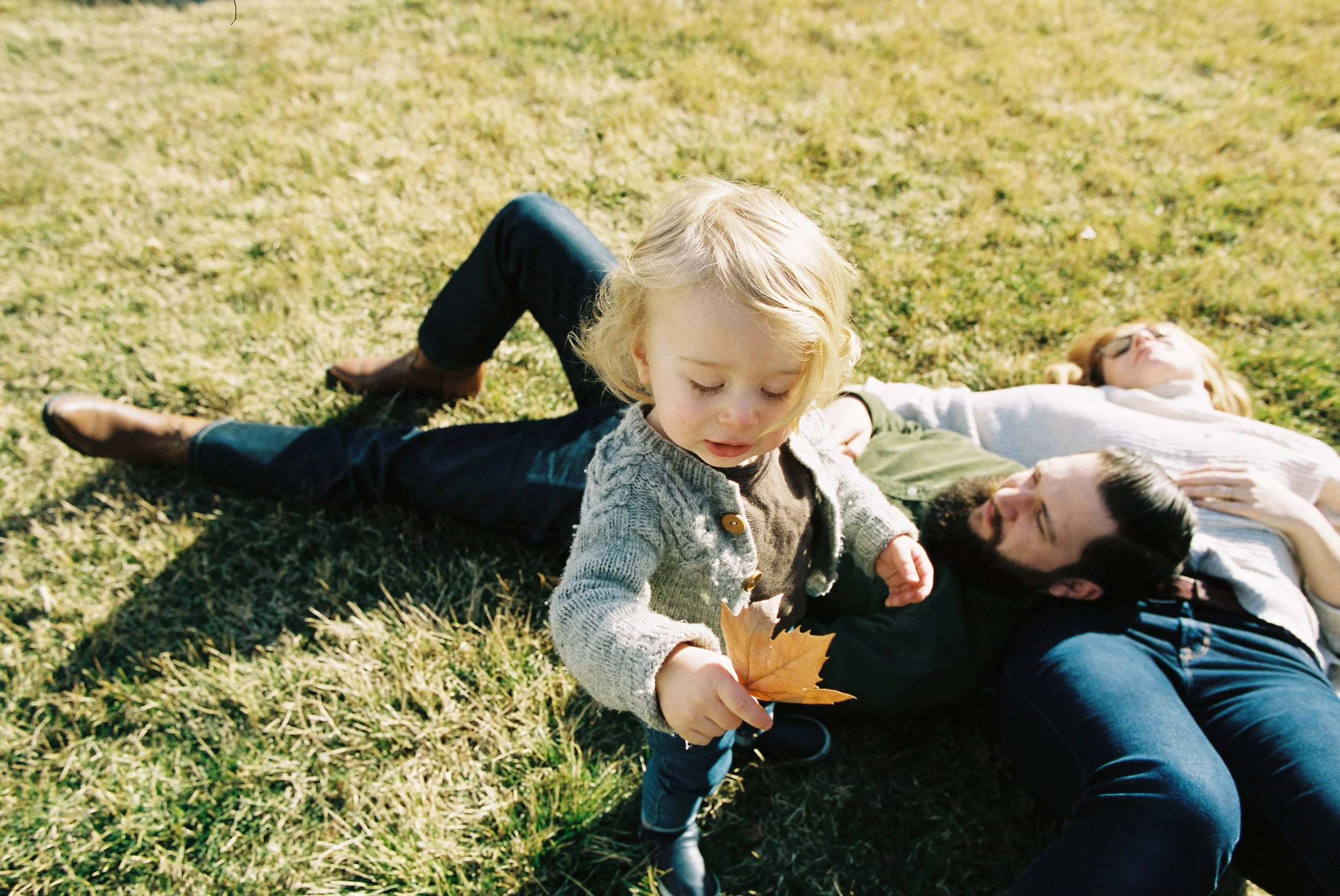 A young girl holding a large autumn leaf while standing on grass, with two adults lying on the ground behind her.