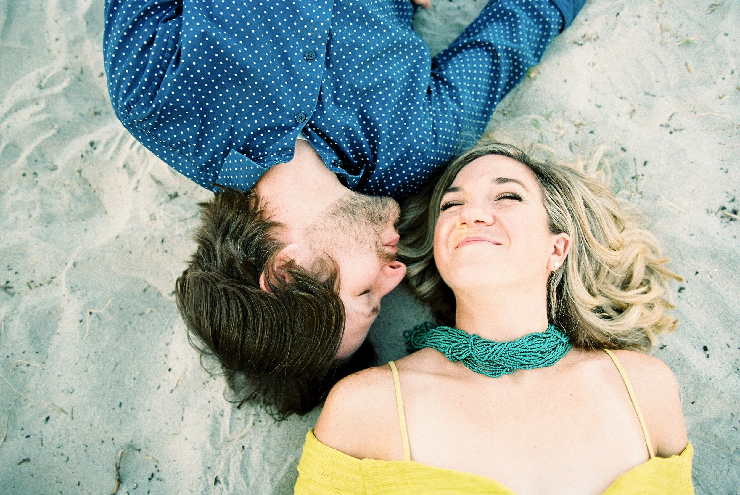 A man and woman lie on the sand, facing each other, smiling and appearing happy.
