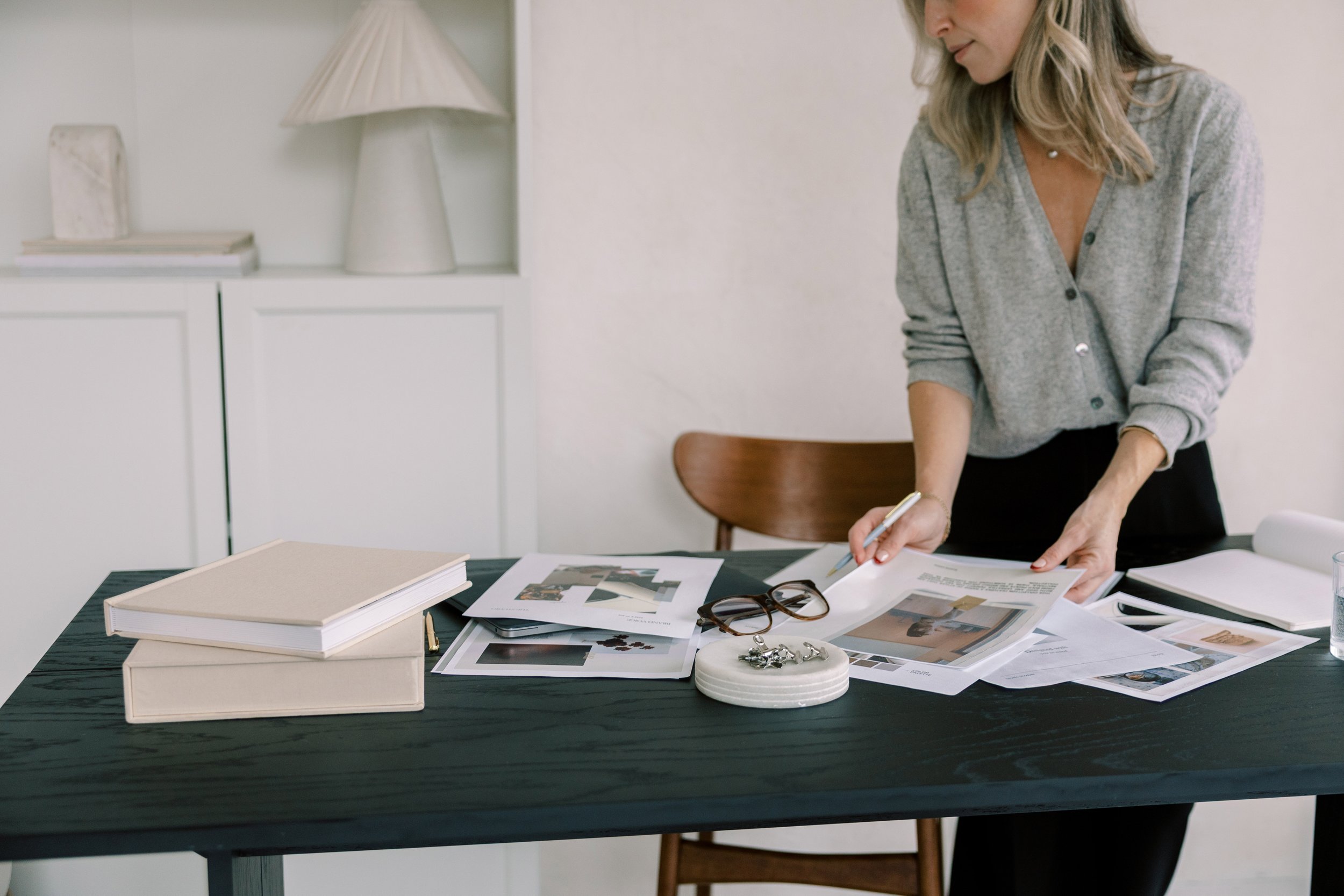 A woman standing at a dark wooden table, looking through papers and photographs, with glasses and jewelry on the table, in a bright room with white cabinets and a wooden chair.