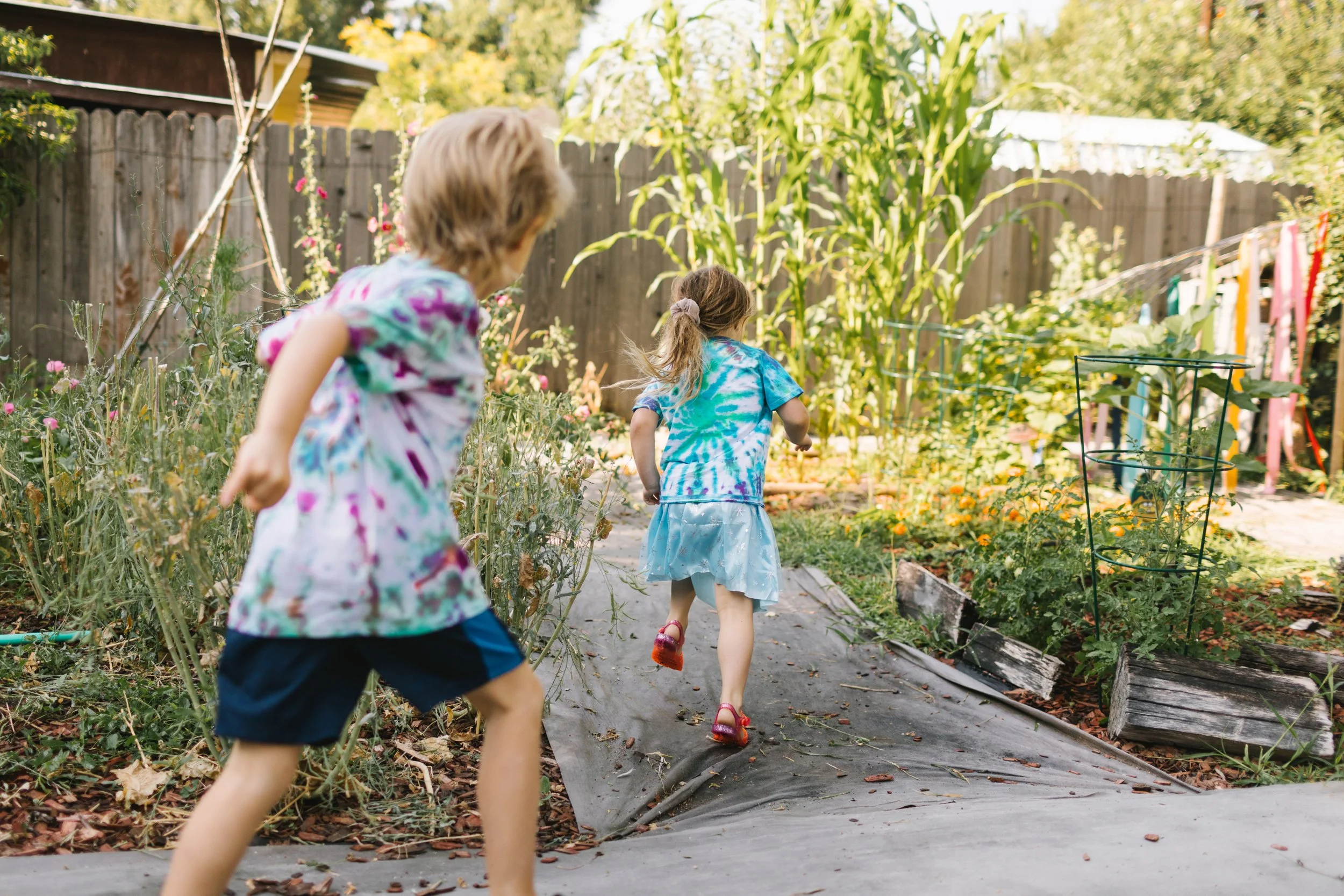 Two children, a boy and a girl, running through a backyard garden with plants and flowers, fenced in with a wooden fence, during daytime.