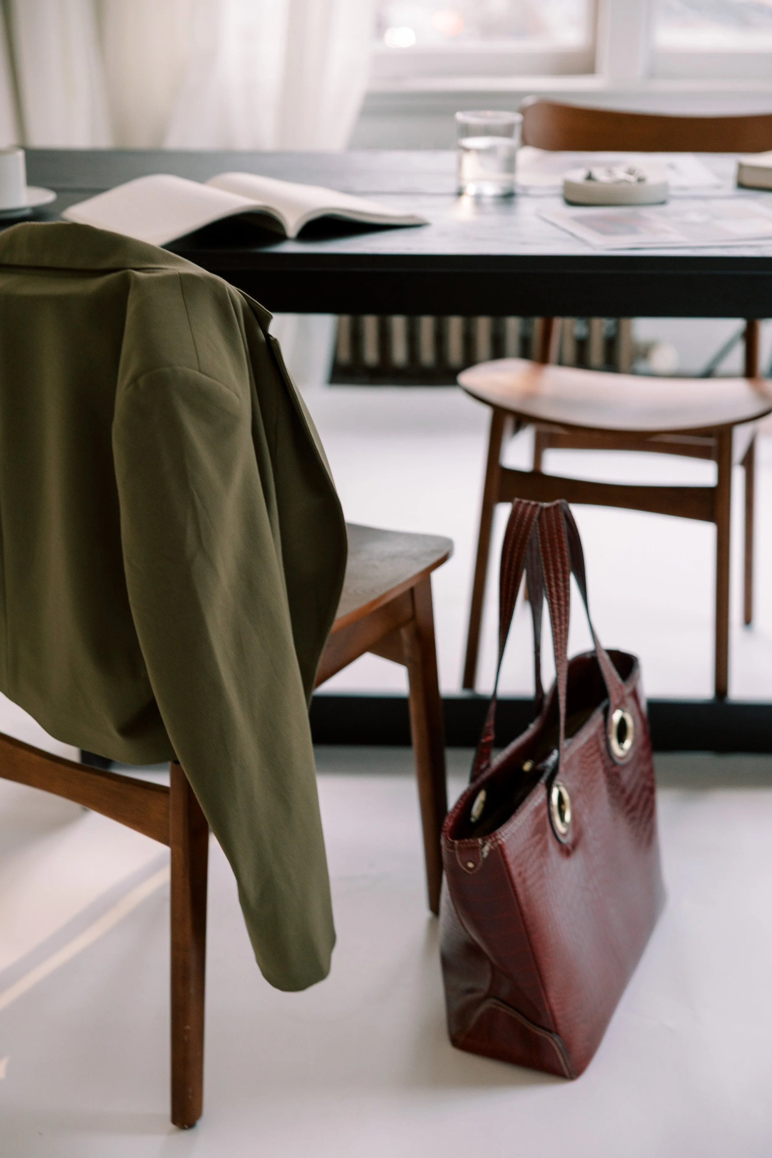 A wooden chair with a green jacket draped over it, a red handbag on the floor, and a table in the background with open books, a glass of water, and magazines.