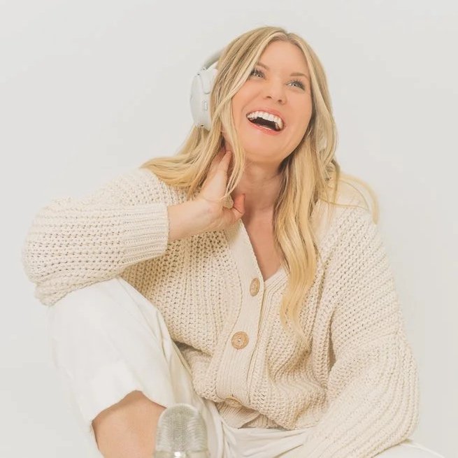 A young woman wearing a beige cardigan and white pants, smiling and singing with her left hand touching her neck, while wearing white headphones against a plain light background.