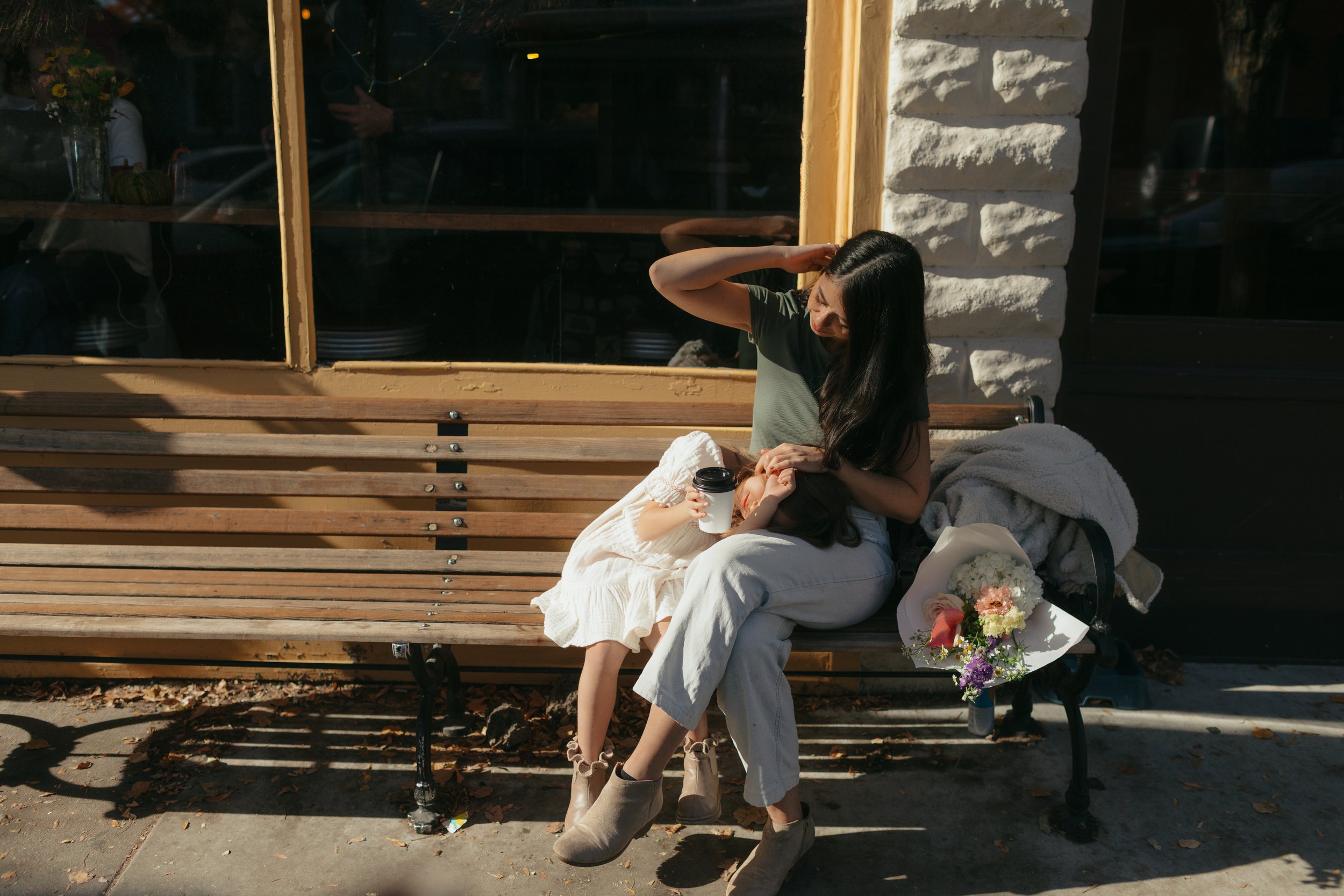 A woman sitting on a wooden bench in Boise, Idaho with a child in a white dress, holding a coffee cup and a flower bouquet, as the woman adjusts the child's hair in a city outdoor setting.