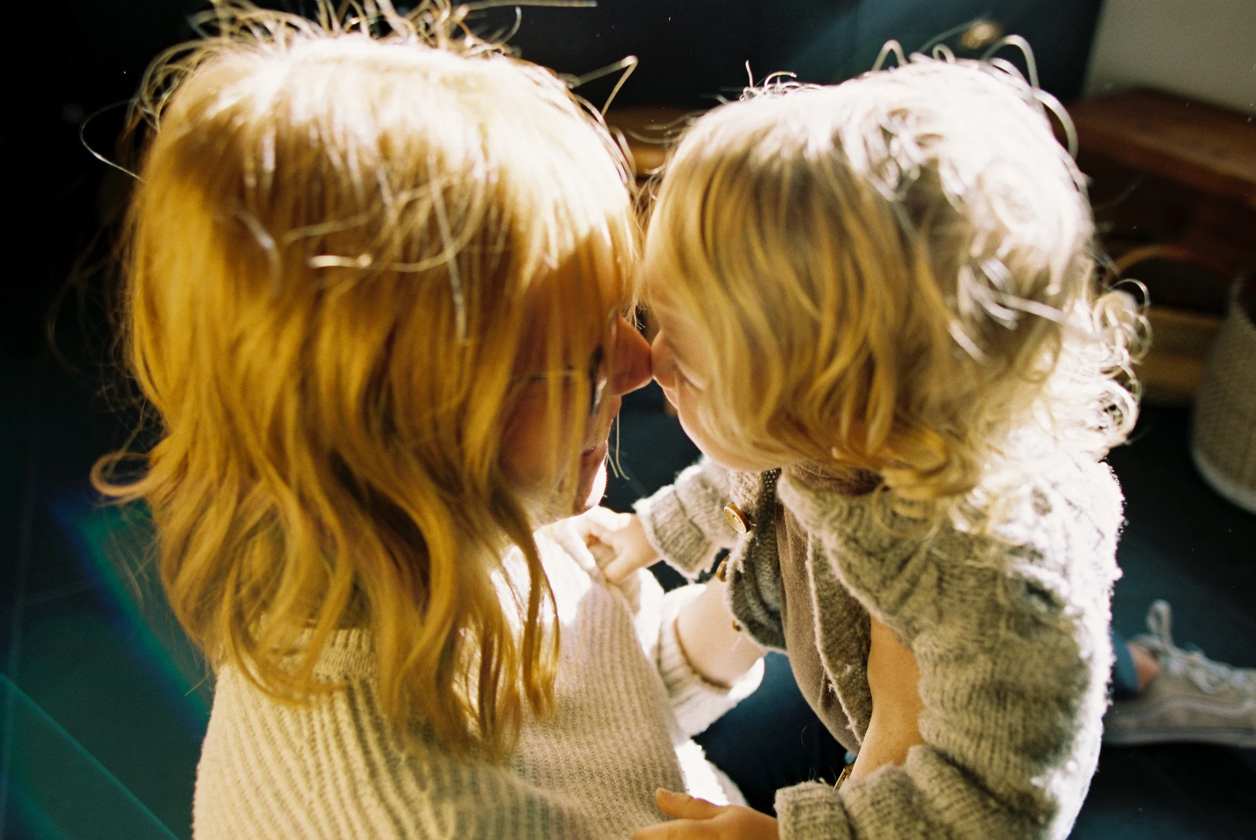 A woman with long, wavy red hair and glasses and a young girl with curly blonde hair touching noses in a close-up indoor shot.  Image taken by Boise Lifestyle family photographer.