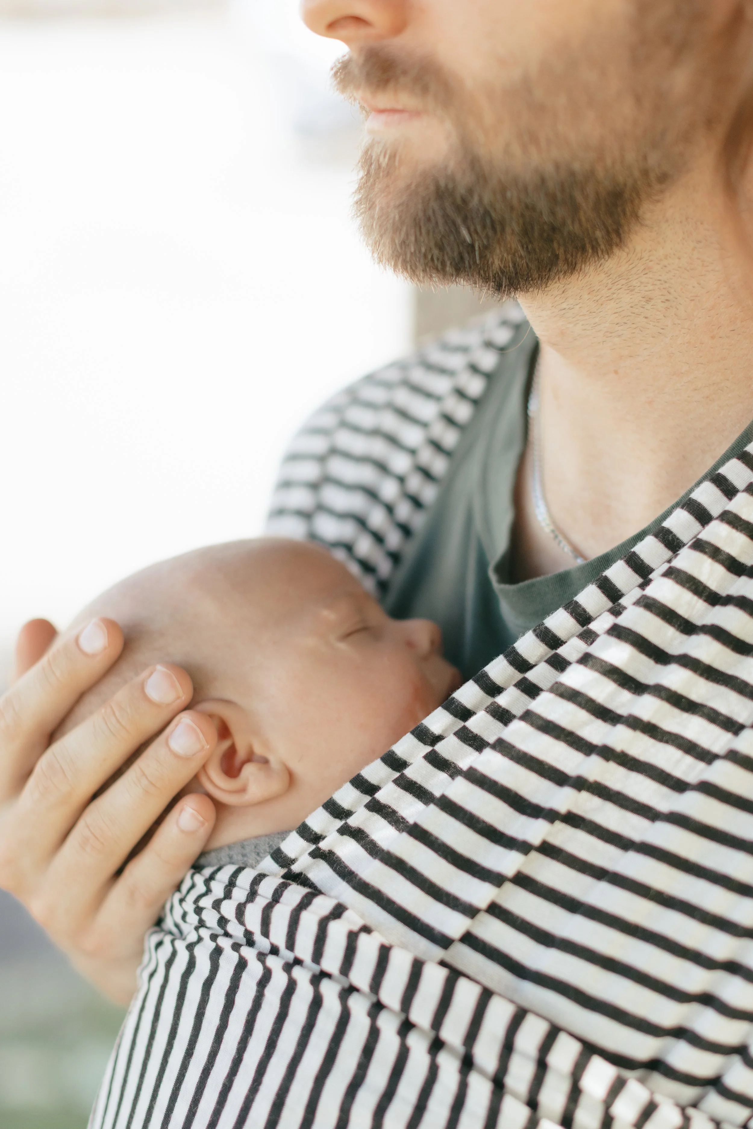 A man with a beard holding a sleeping baby close to his chest.