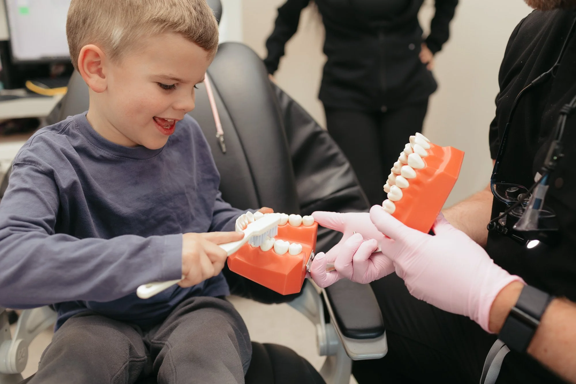 A young boy smiling while practicing brushing teeth on a dental model, held by a person wearing gloves, during a dental health demonstration.
