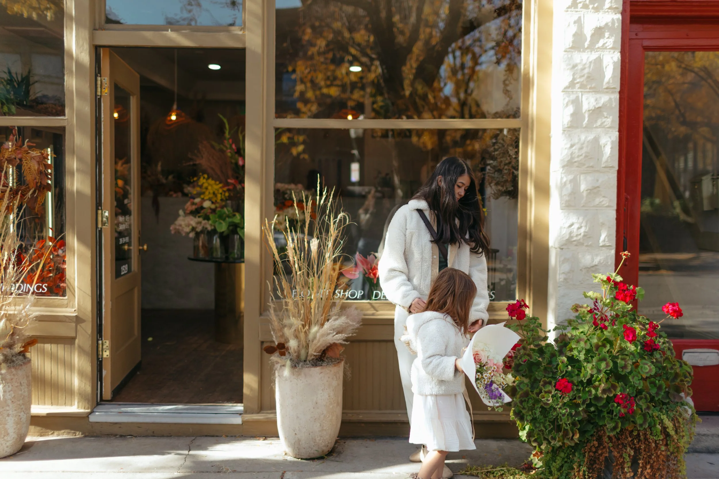 A woman and a young girl stand outside a flower shop in Boise, Idaho, looking at a bouquet, with autumn leaves visible through the shop window.