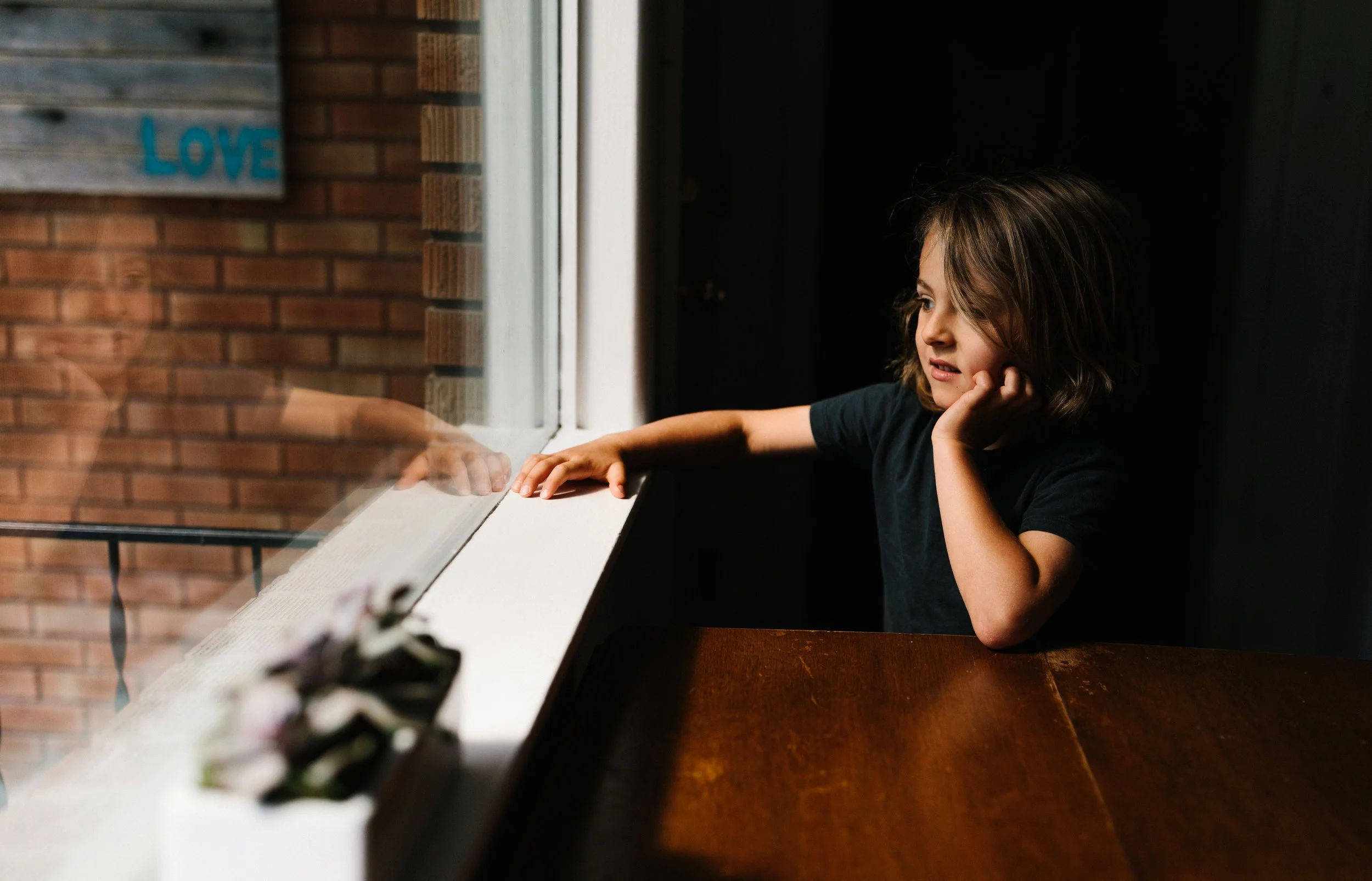 A young boy with shoulder-length hair, sitting at a dinner table by a window, looking outside with a bored or contemplative expression, hand resting on his cheek, and reaching out to touch a book or magazine on the windowsill.