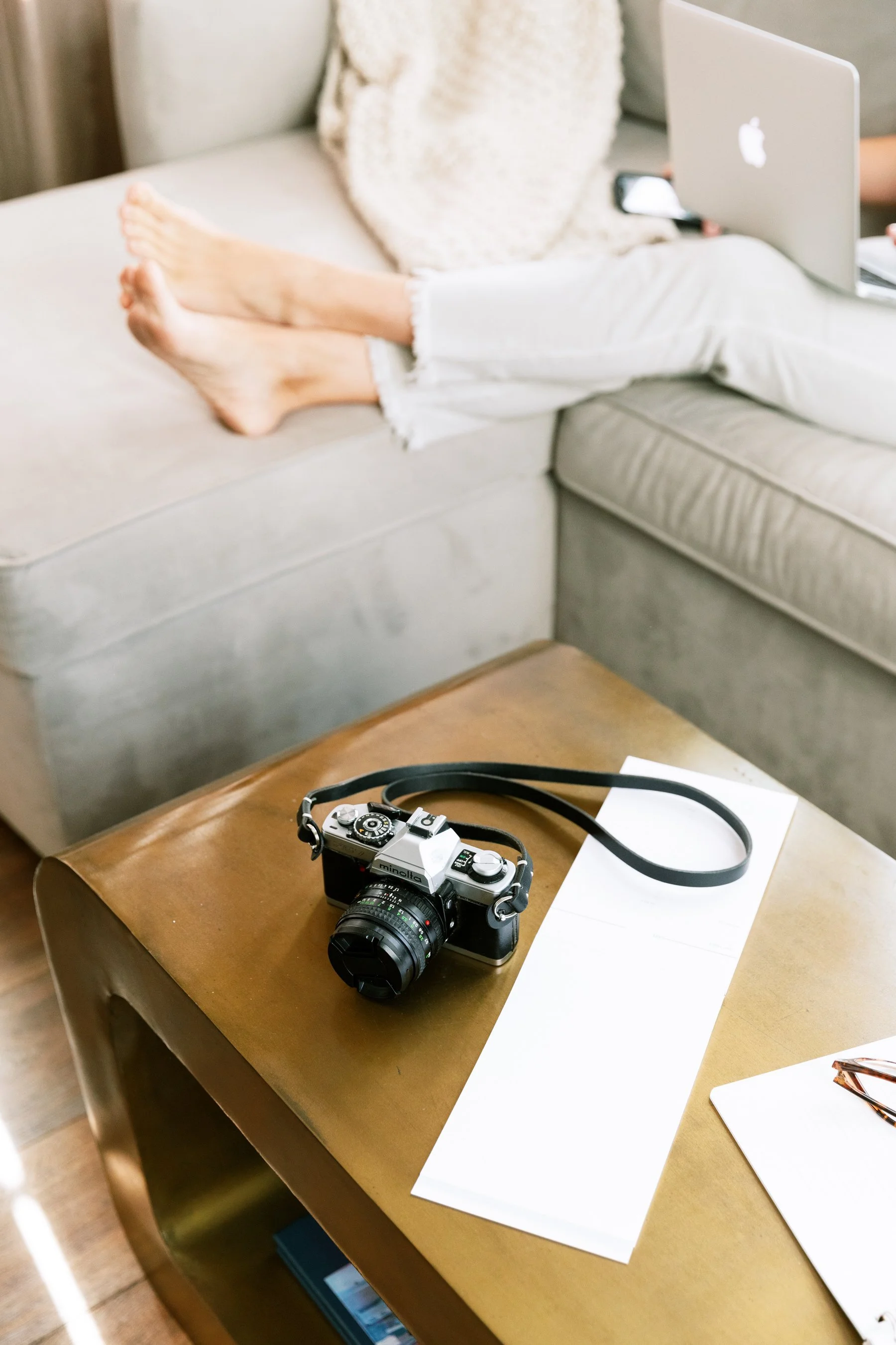 A vintage camera resting on a wooden coffee table, with a white paper and a pair of glasses nearby, in front of a sofa where a person is sitting with legs extended, using a laptop.