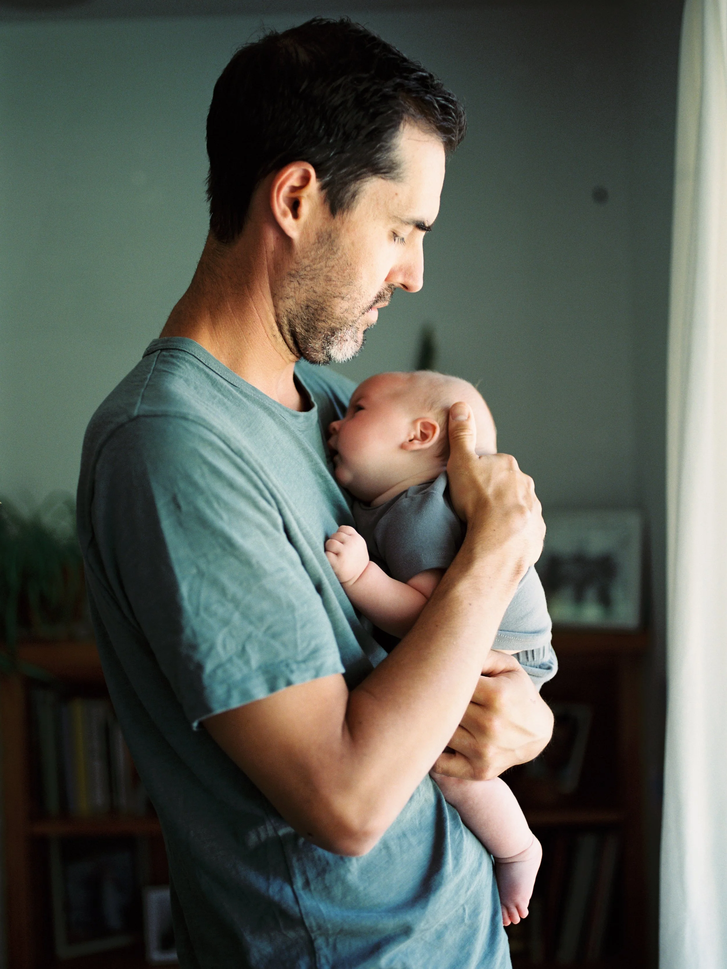A man holding a baby close to his chest, with his face gently touching the baby's head, in a cozy indoor setting with a bookshelf in the background.