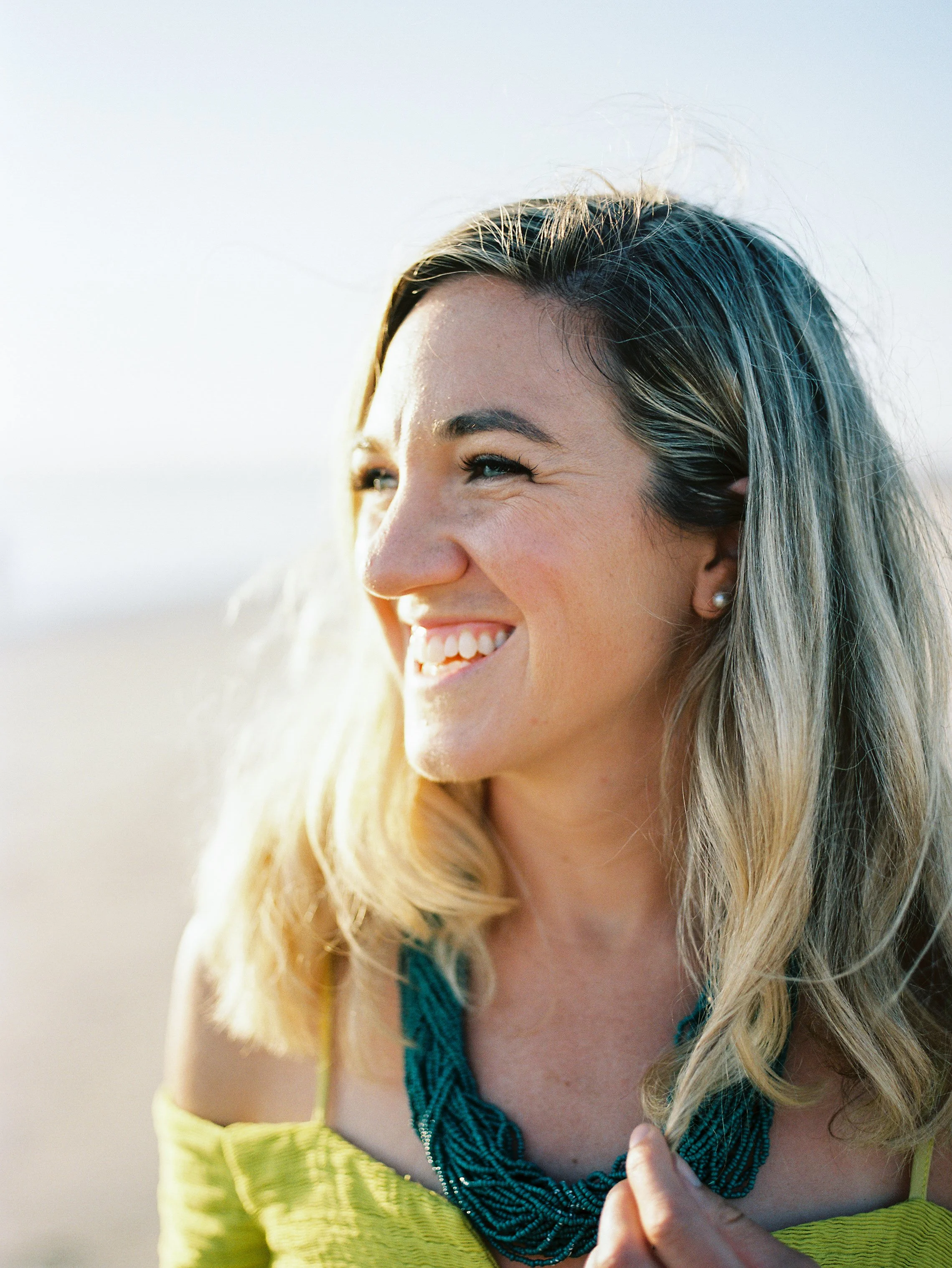 Close-up of a smiling woman with long blonde hair, wearing a yellow top and a blue beaded necklace, outdoors near the beach.