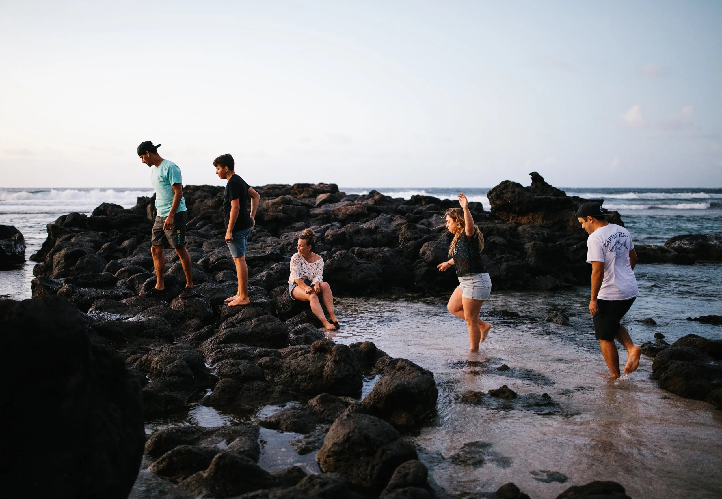 Group of people exploring rocks and shallow water at the beach during sunset.