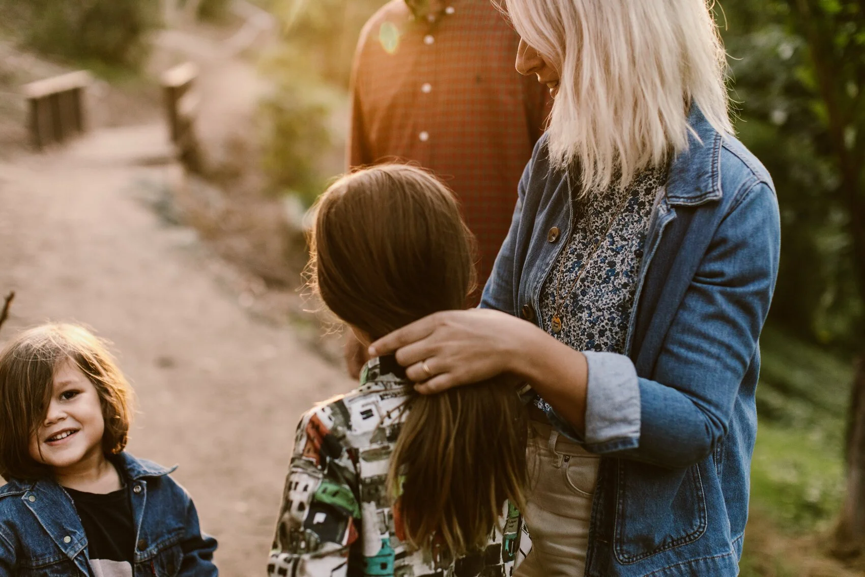 A woman with blonde hair wearing a denim jacket smiling and gently holding a young girl with brown hair. Two other children, also with brown hair, are nearby outdoors on a pathway surrounded by greenery.