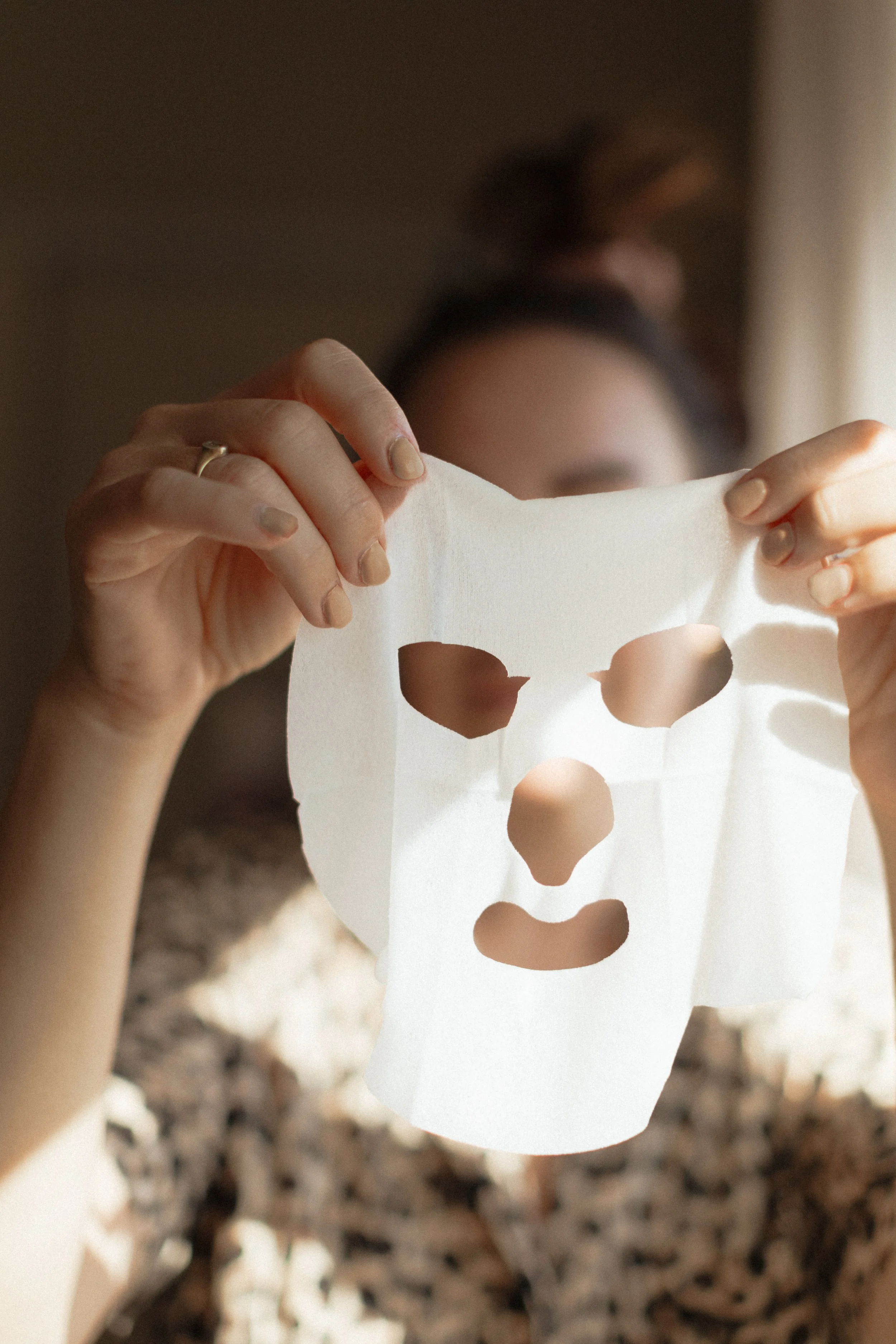 Person holding a white sheet mask in front of their face, with the mask’s eye, nose, and mouth holes visible.
