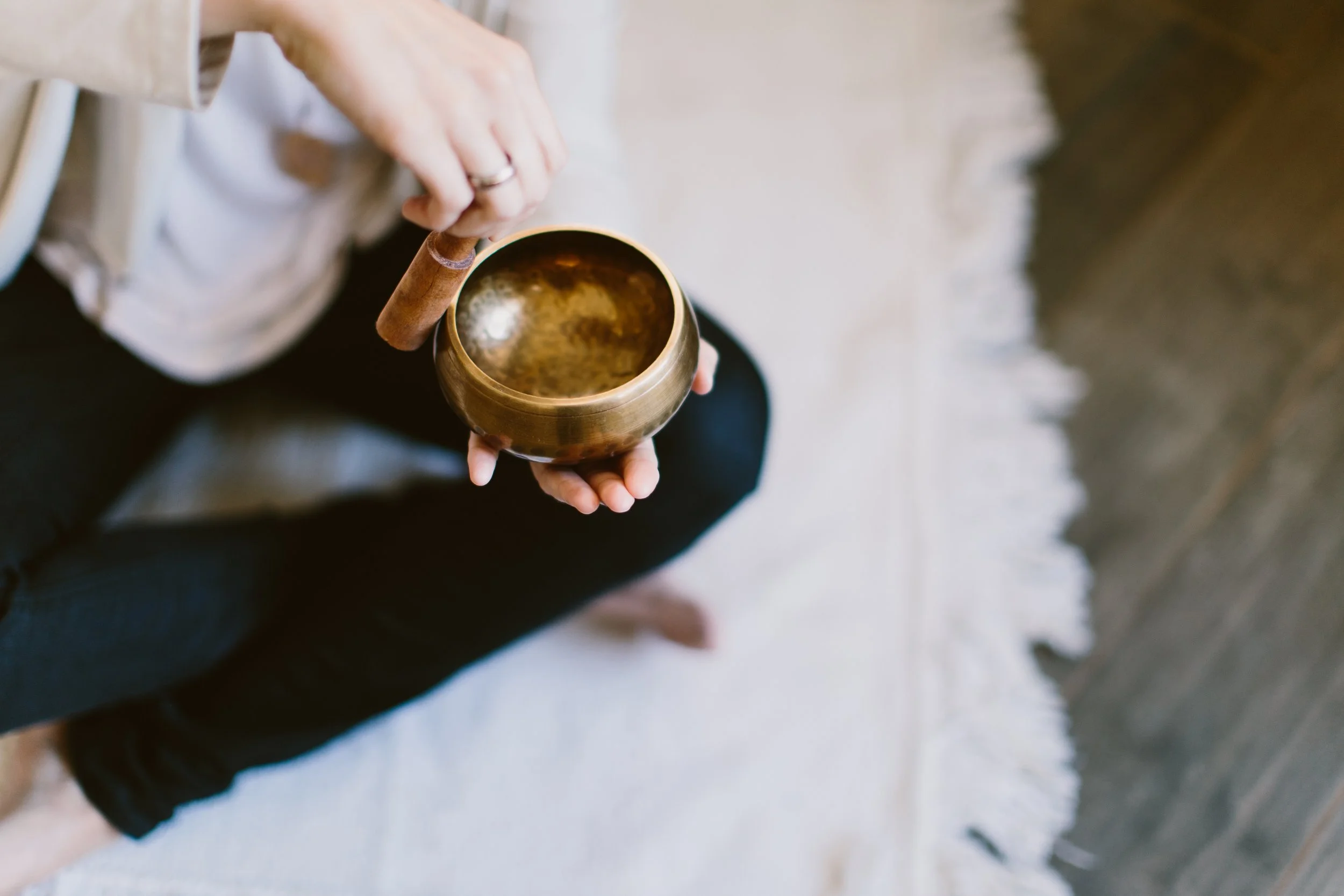 Person holding a singing bowl and a mallet, sitting on a white rug near a wooden floor