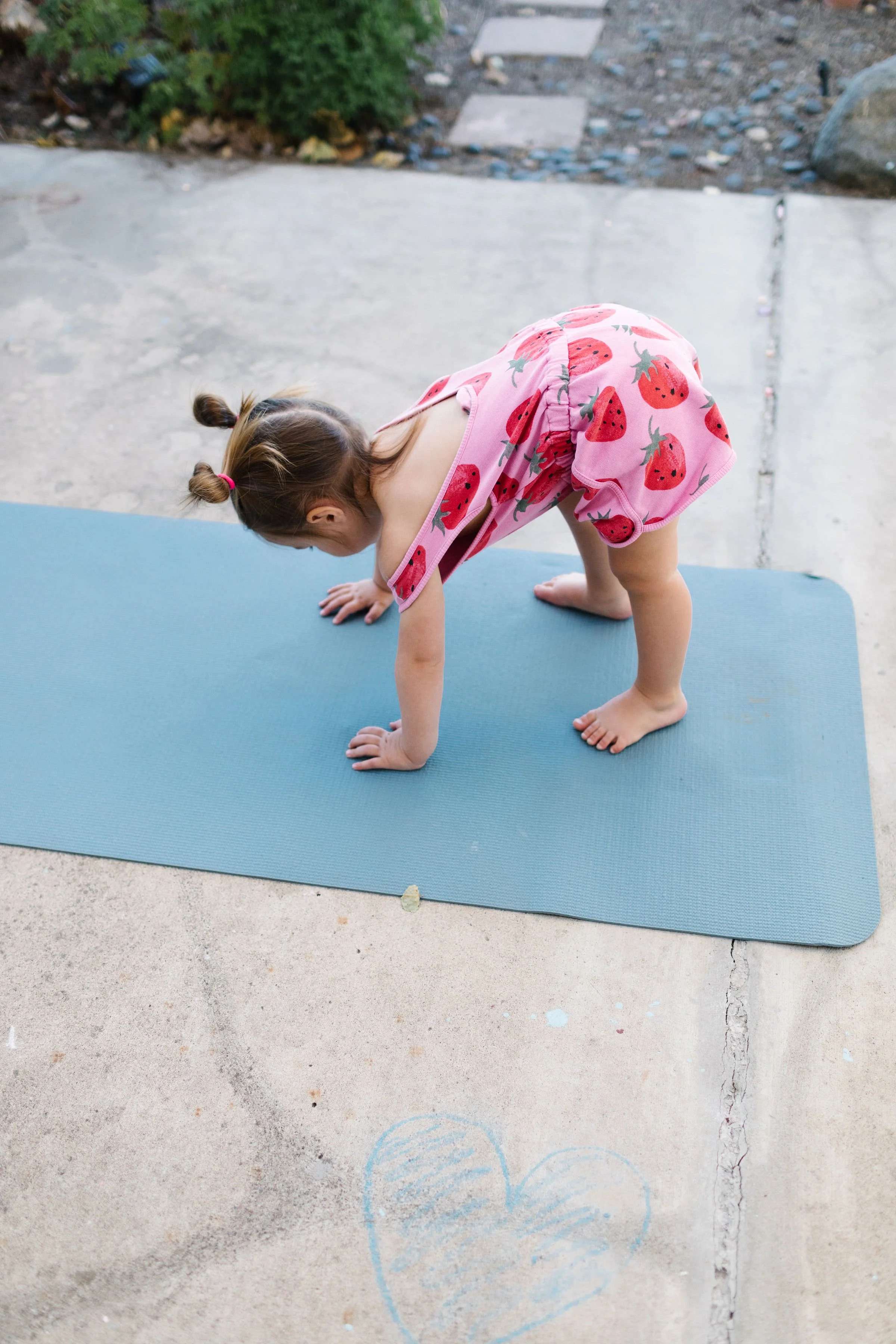 A young girl in a pink strawberry-patterned dress is bending forward, placing her hands and feet on a blue exercise mat on a concrete porch, outdoors.