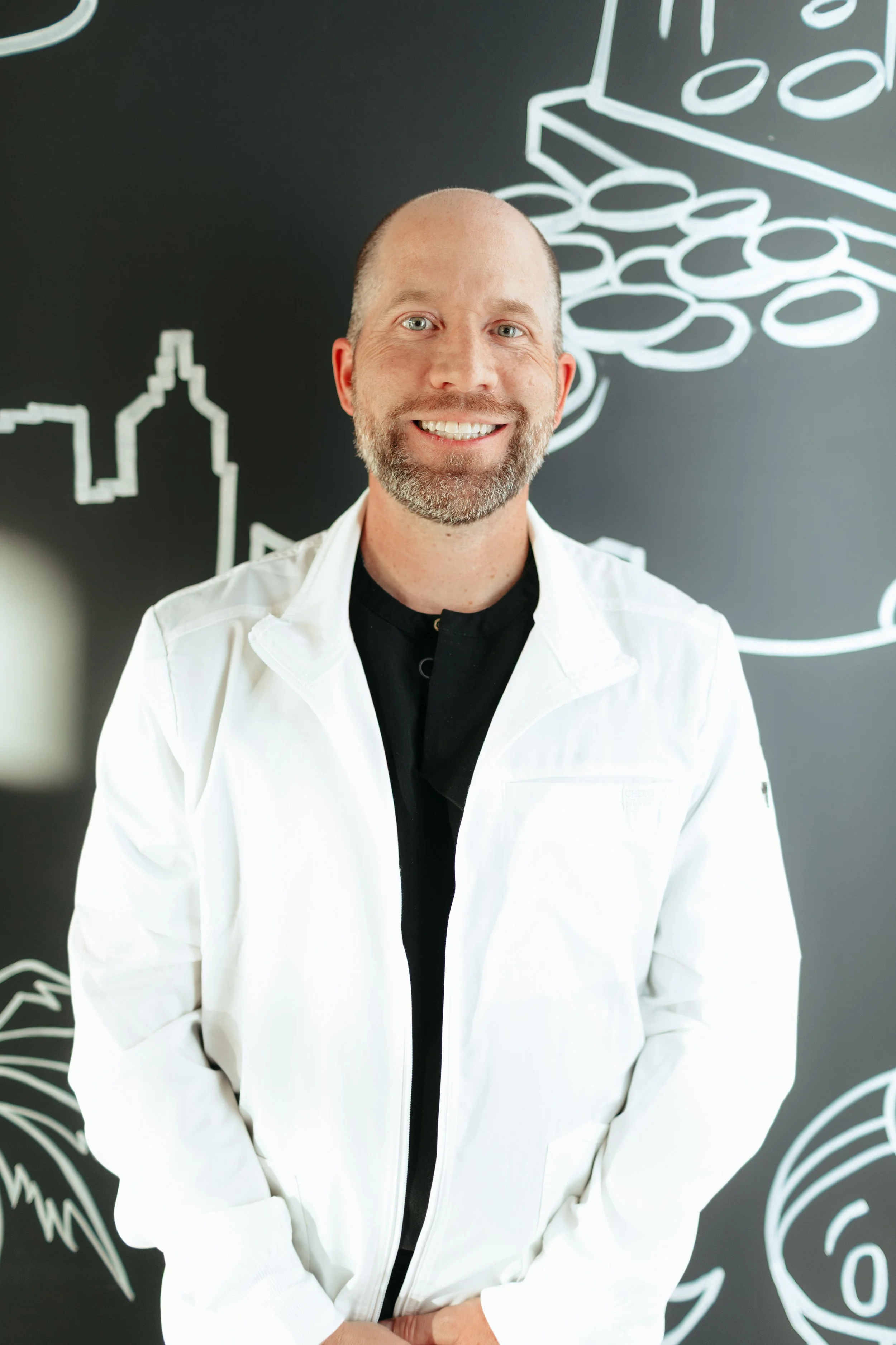 A smiling dentist wearing a white jacket, standing in front of a blackboard with white chalk drawings of food and a city skyline.
