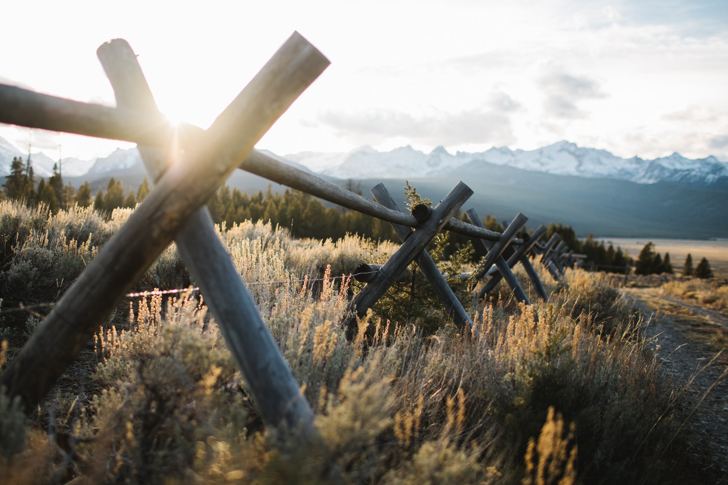 Wooden fence on a trail in a field with mountains in the background, during sunset or sunrise.