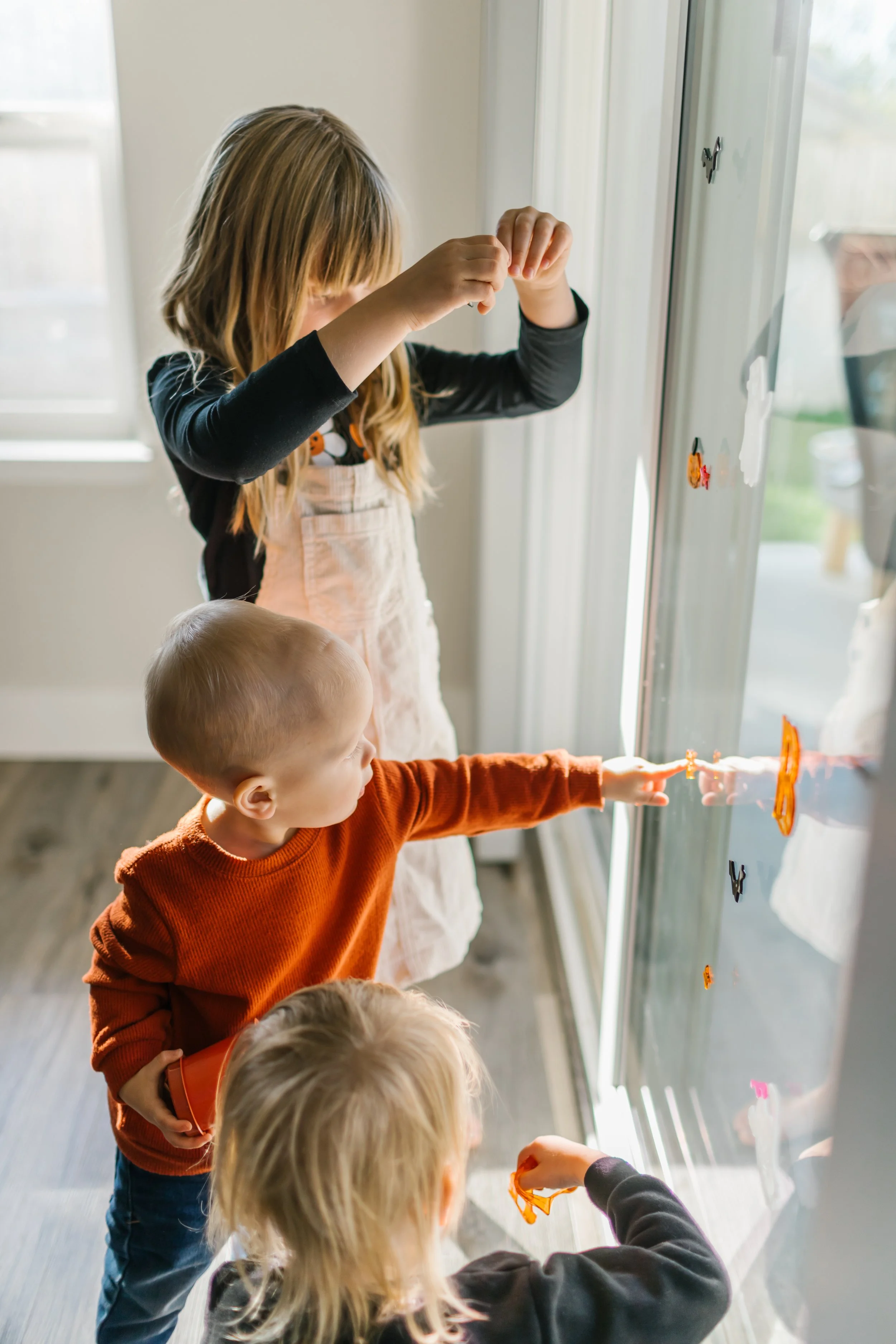 Three children, a girl and two boys, are decorating a glass door with colorful stickers indoors, near a window.