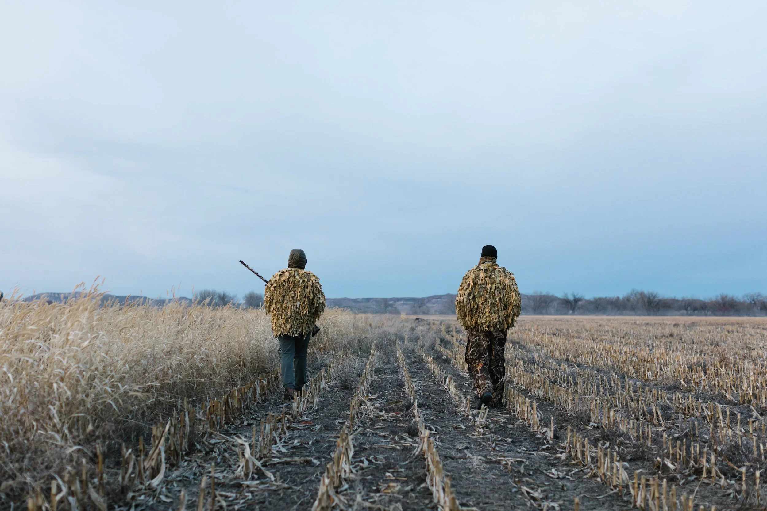 Two hunters in camouflage jackets walking through a harvested cornfield on a cloudy day, with one carrying a rifle.