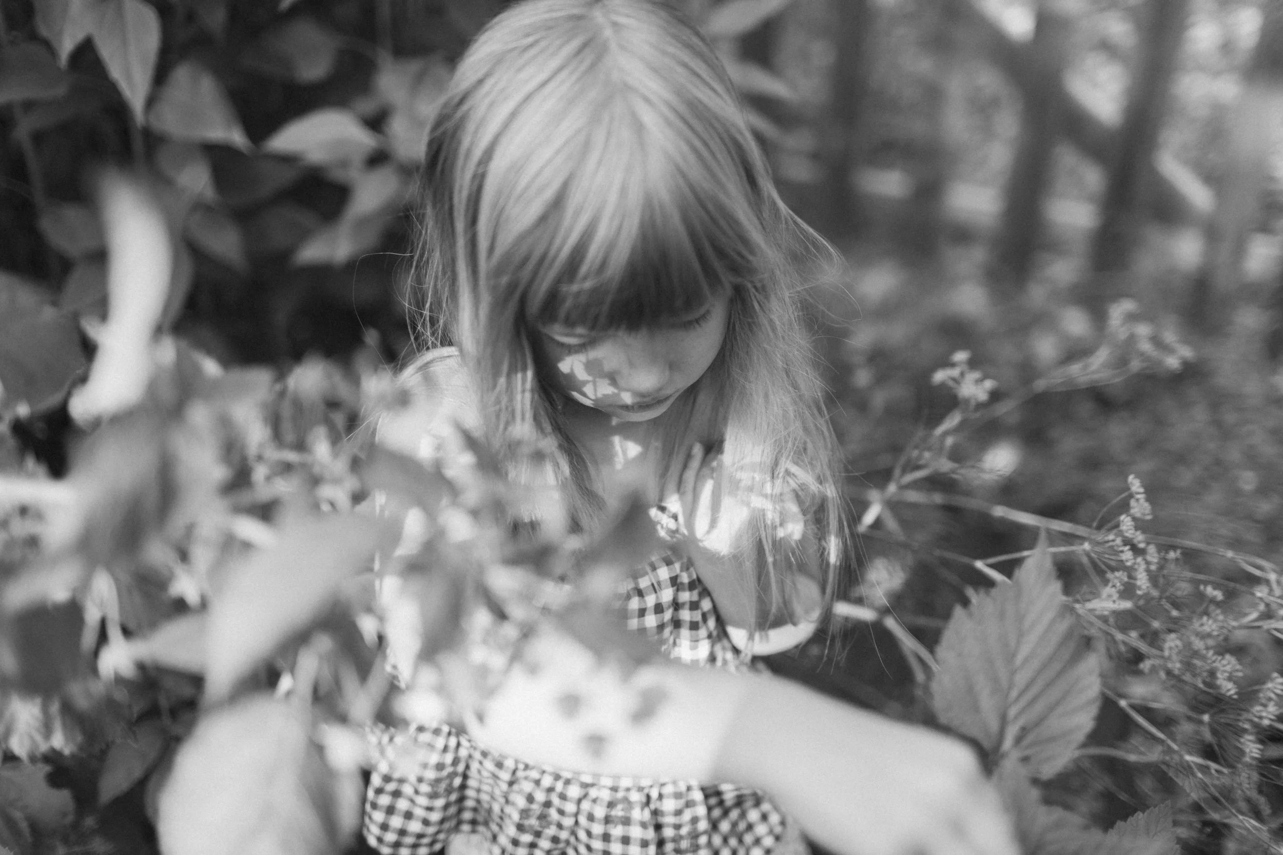 Black and white photo of a young girl with long hair and face paint, looking down and surrounded by foliage, outdoors in a natural setting.