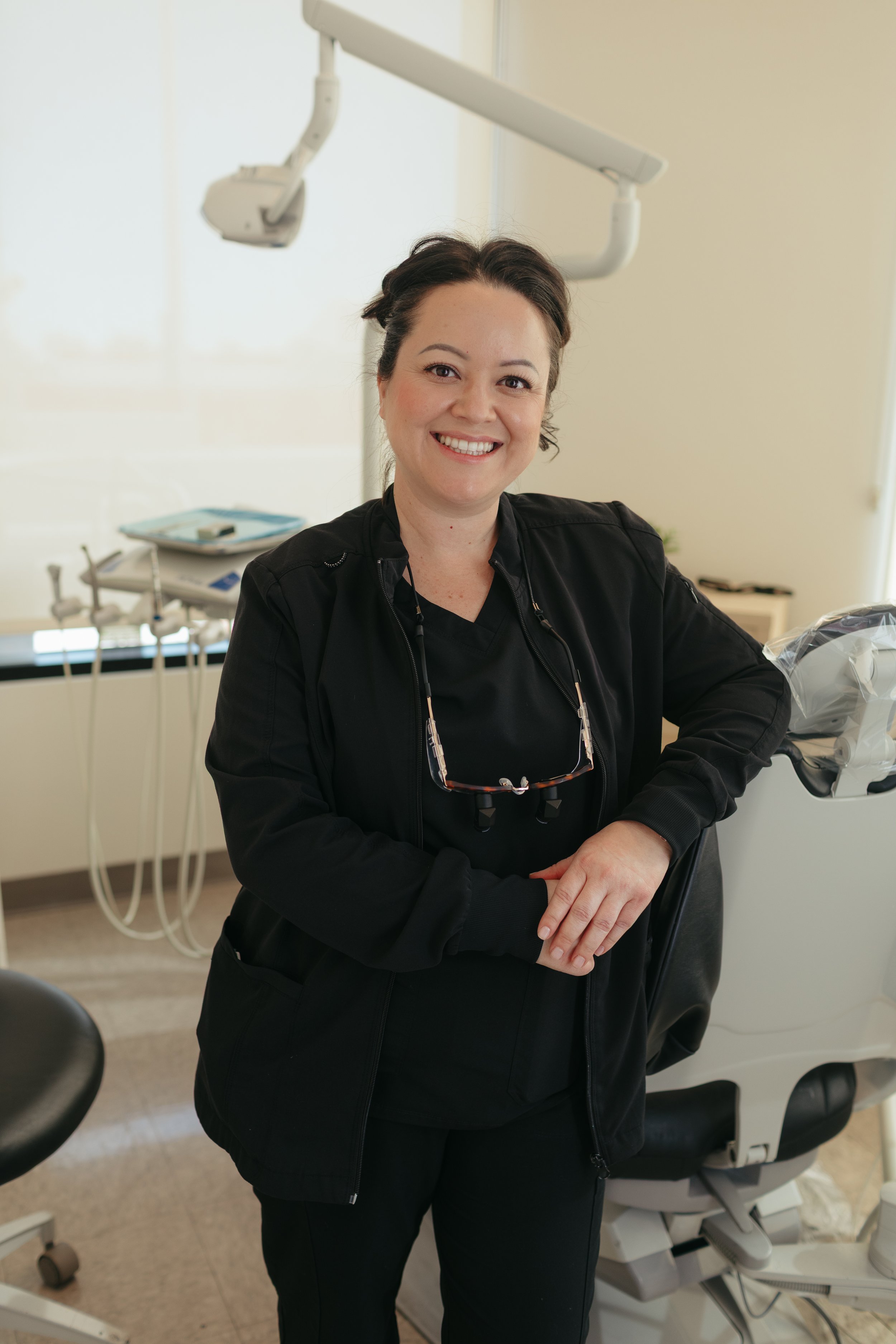 A smiling woman in black scrubs and glasses hanging around her neck in a dental office, standing next to a dental chair and equipment.