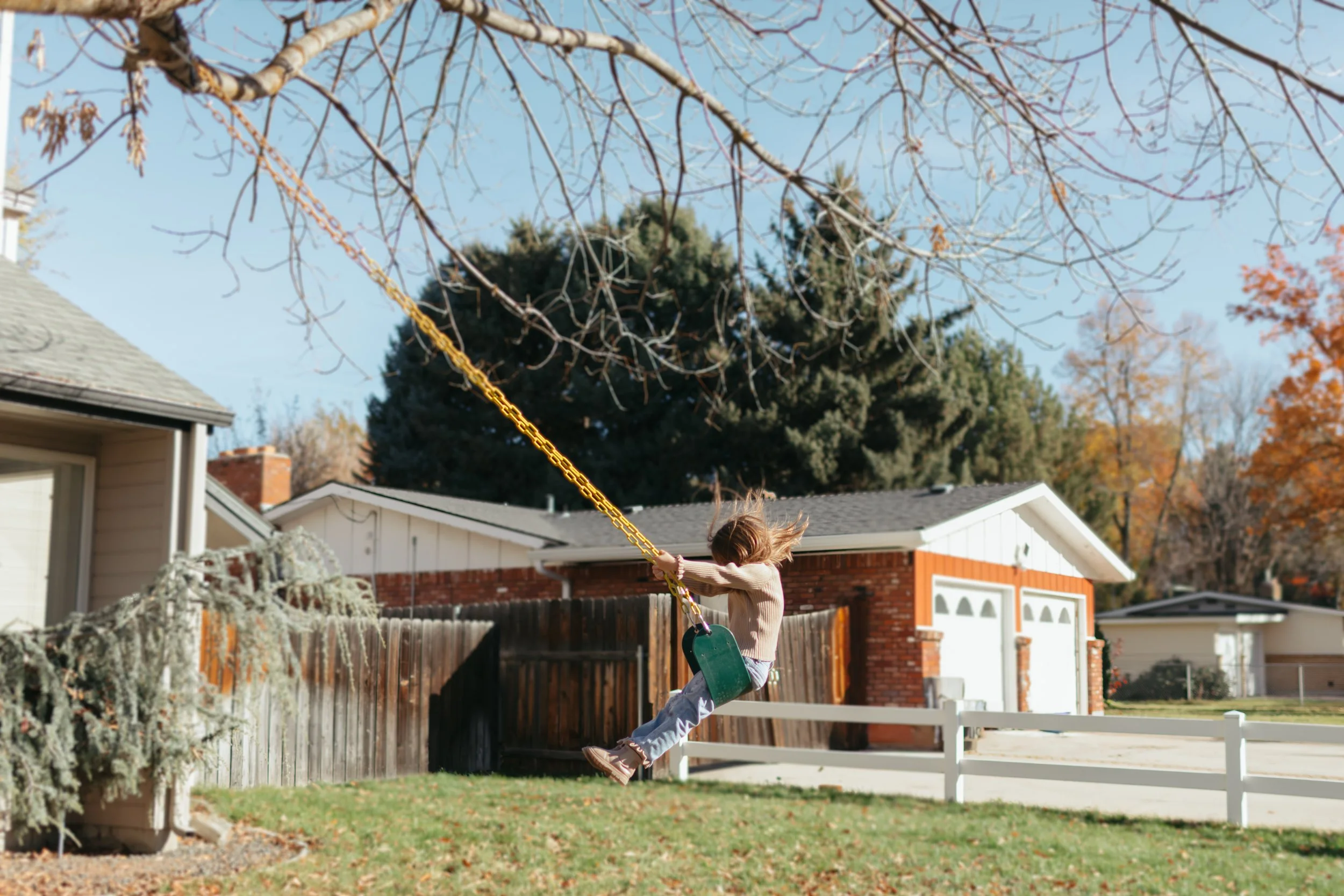 A young girl swinging on a backyard swing set on a sunny autumn day. The girl is holding onto the chains of the swing, which are yellow, and she is mid-air with her hair flying. The yard is surrounded by a wooden fence with trees and houses in the ba