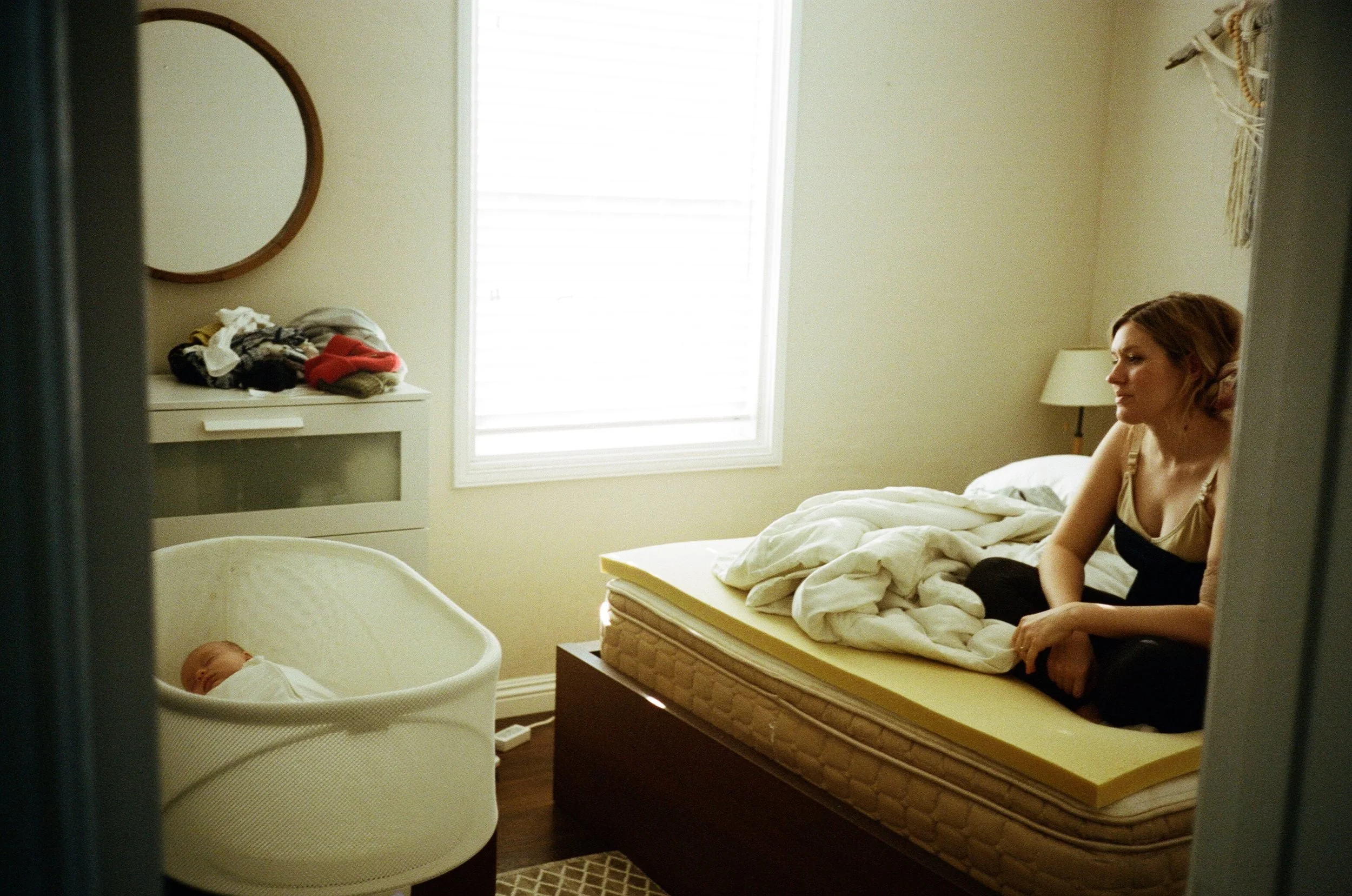 A woman sitting on a bed in a bedroom, next to a bassinet with a sleeping baby. The room has light-colored walls, a window with white blinds, a small table with a lamp, and a dresser with laundry on top.