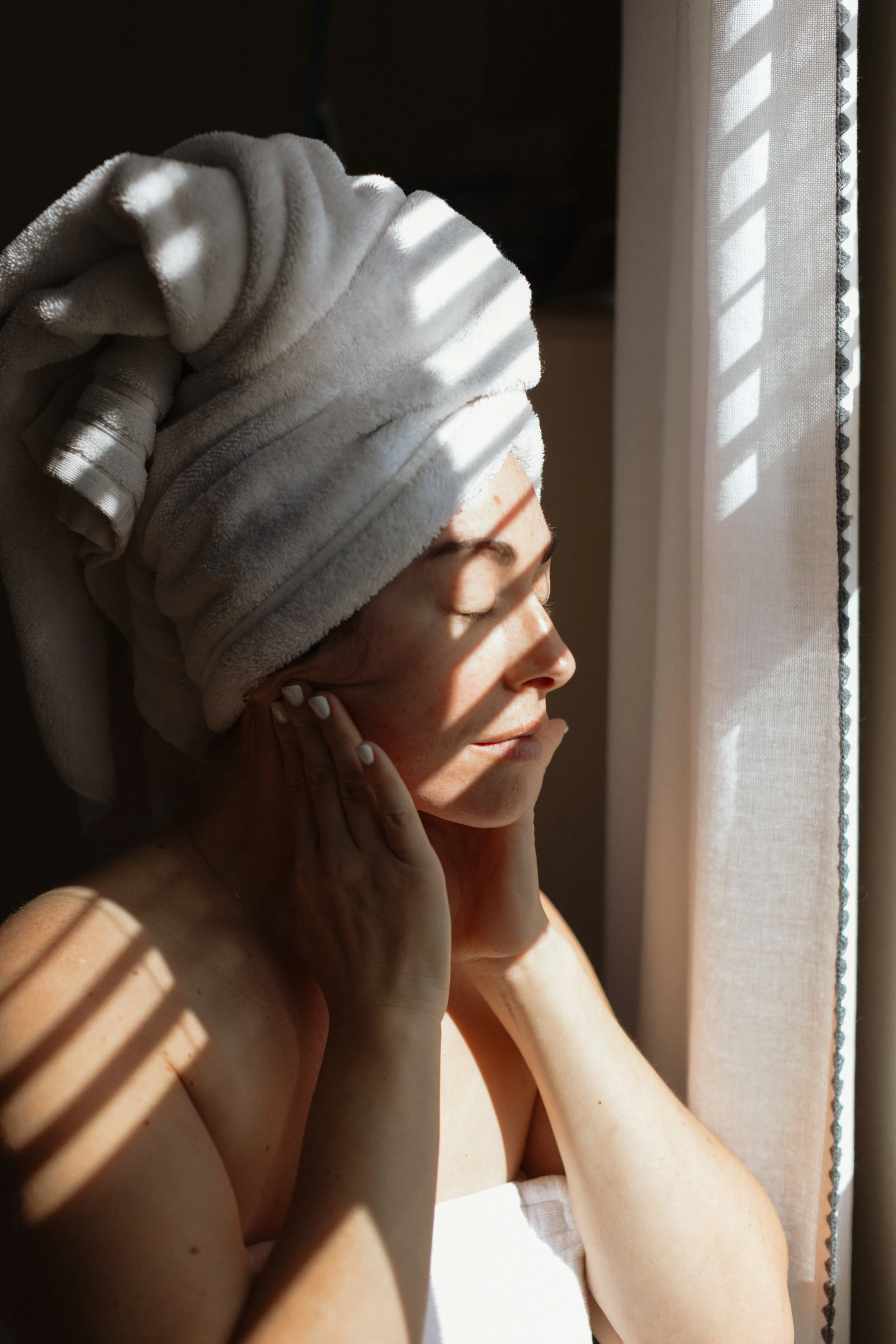 A woman with a towel wrapped around her head, standing near a window with sunlight casting shadows through blinds, listening to music with headphones.