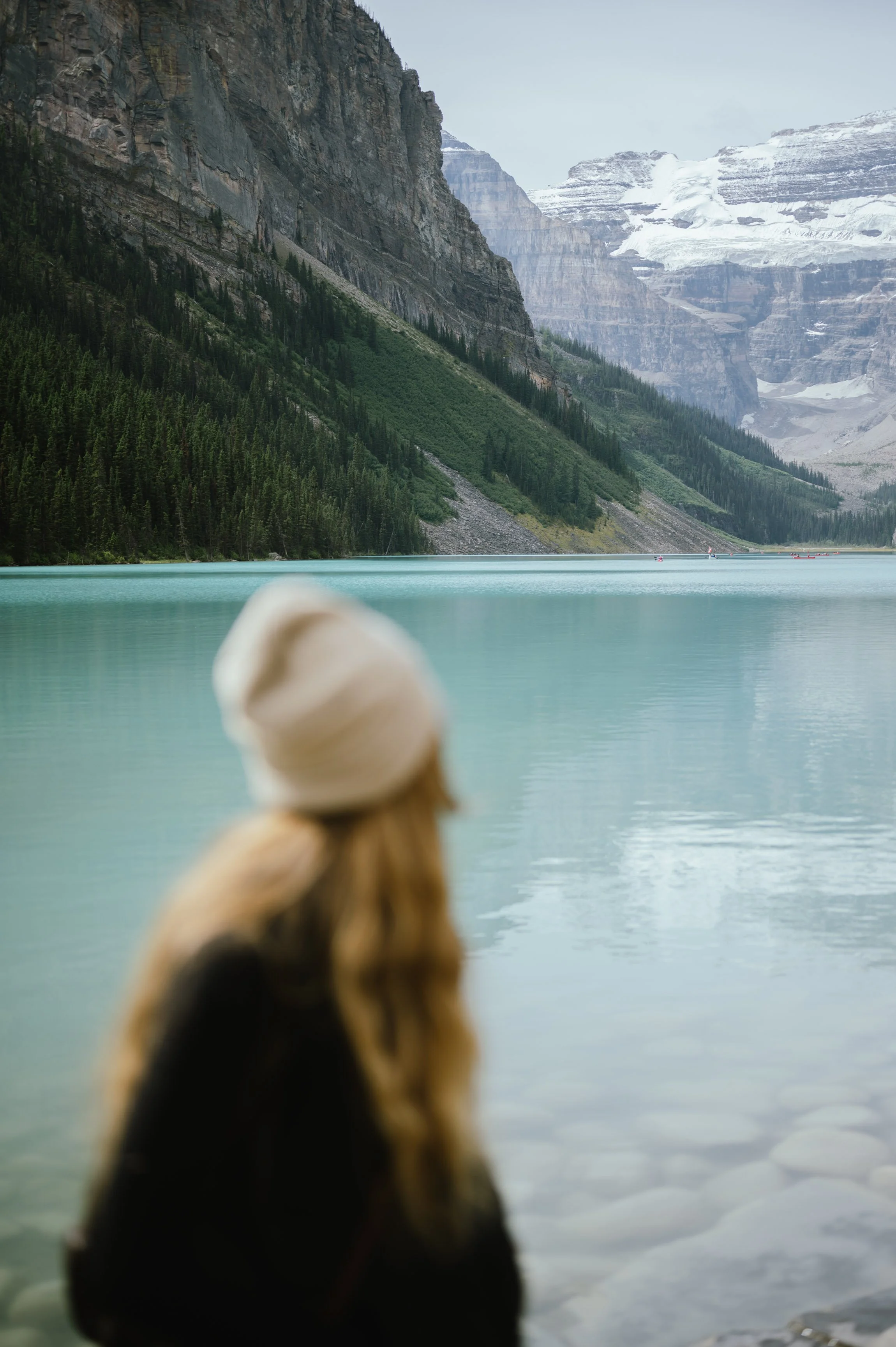 A woman with long hair, wearing a white beanie and dark jacket, sitting by a lake with mountain and forest scenery in the background.