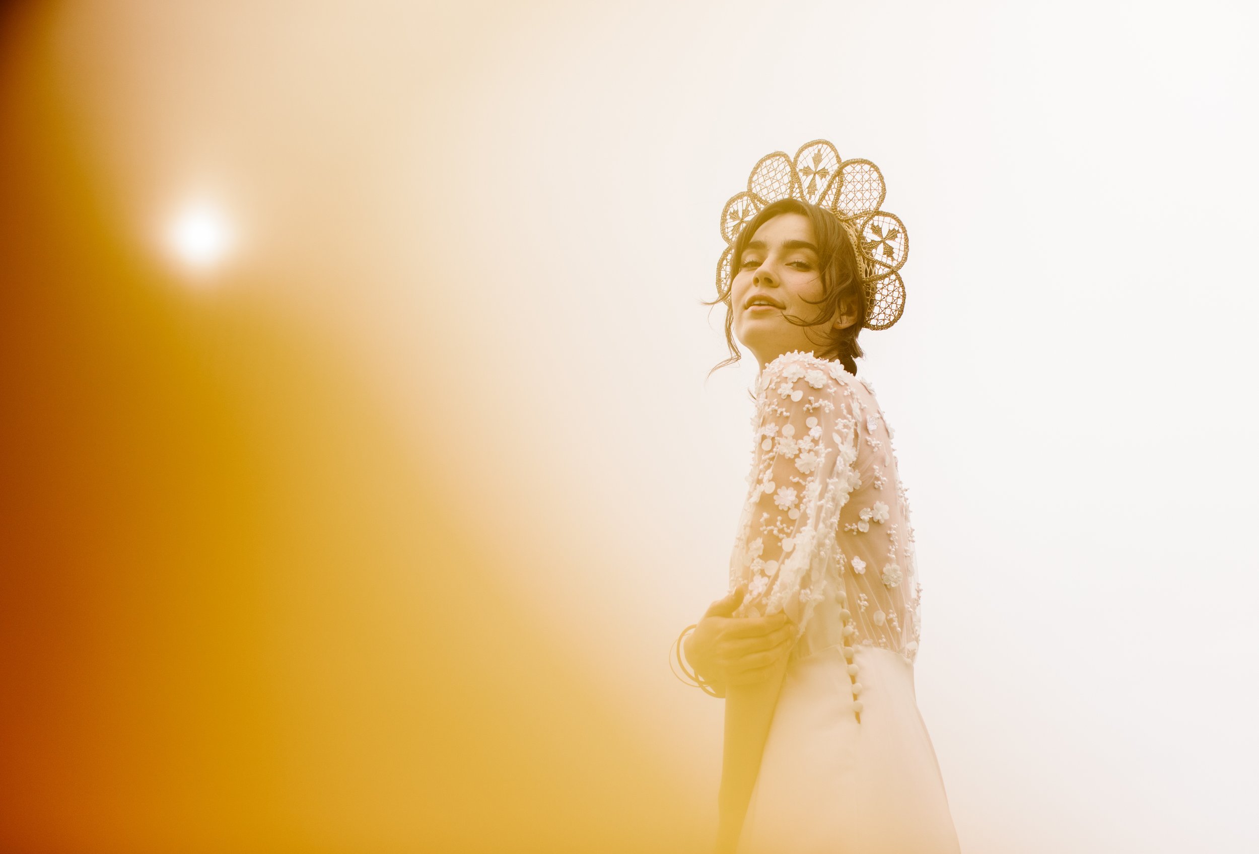A young woman wearing a white dress with floral embellishments and a lace neckline, standing outdoors against a bright sky with a decorative headpiece made of woven circles.