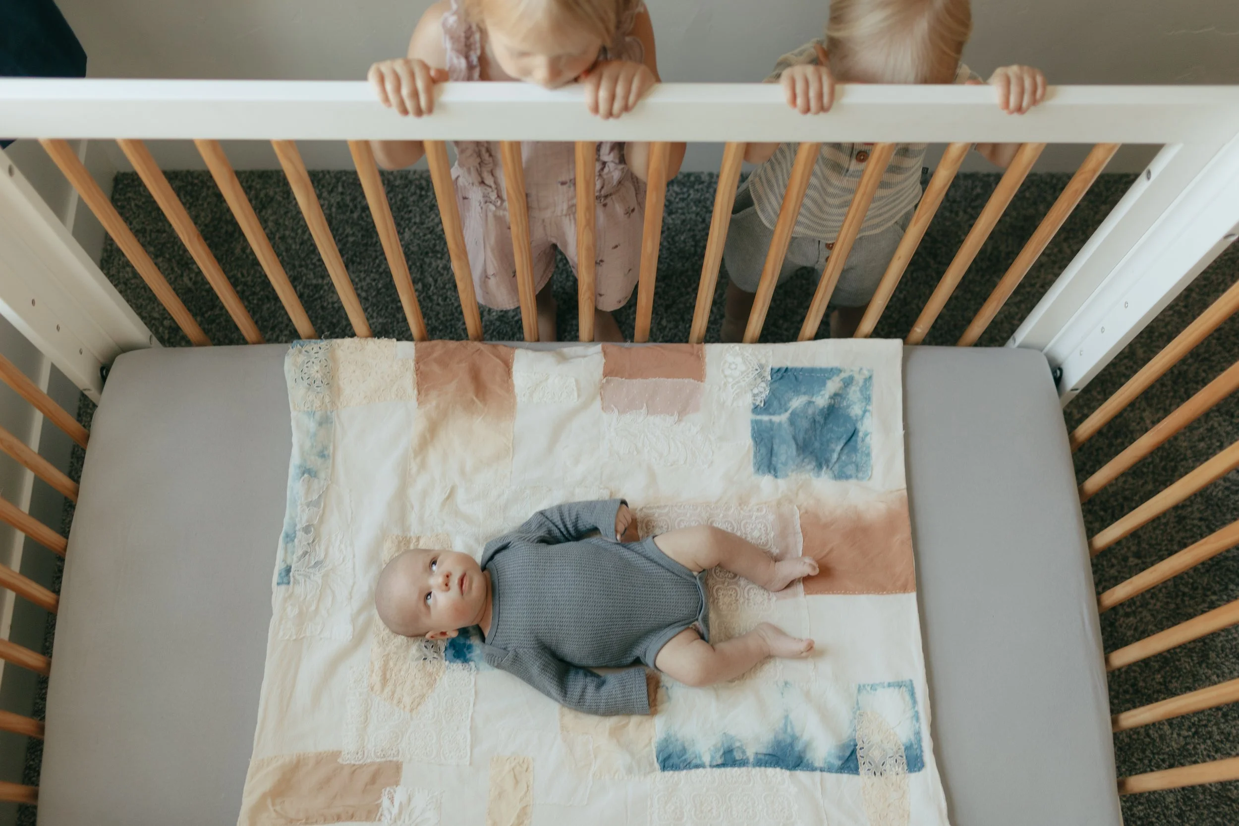 A baby lying on a quilt in a crib, looking up, with two children peering over the side of the crib and looking down at the baby.