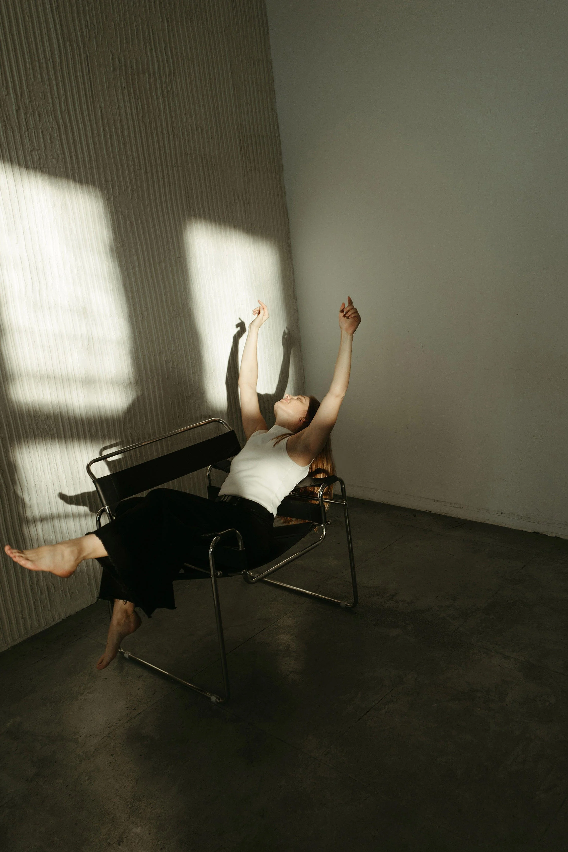 A woman in a white top and black pants is sitting on a black bench with metal arms, leaning back with her arms raised and her eyes closed. Shadows of her hands are cast on a textured wall, as sunlight streams through a window.