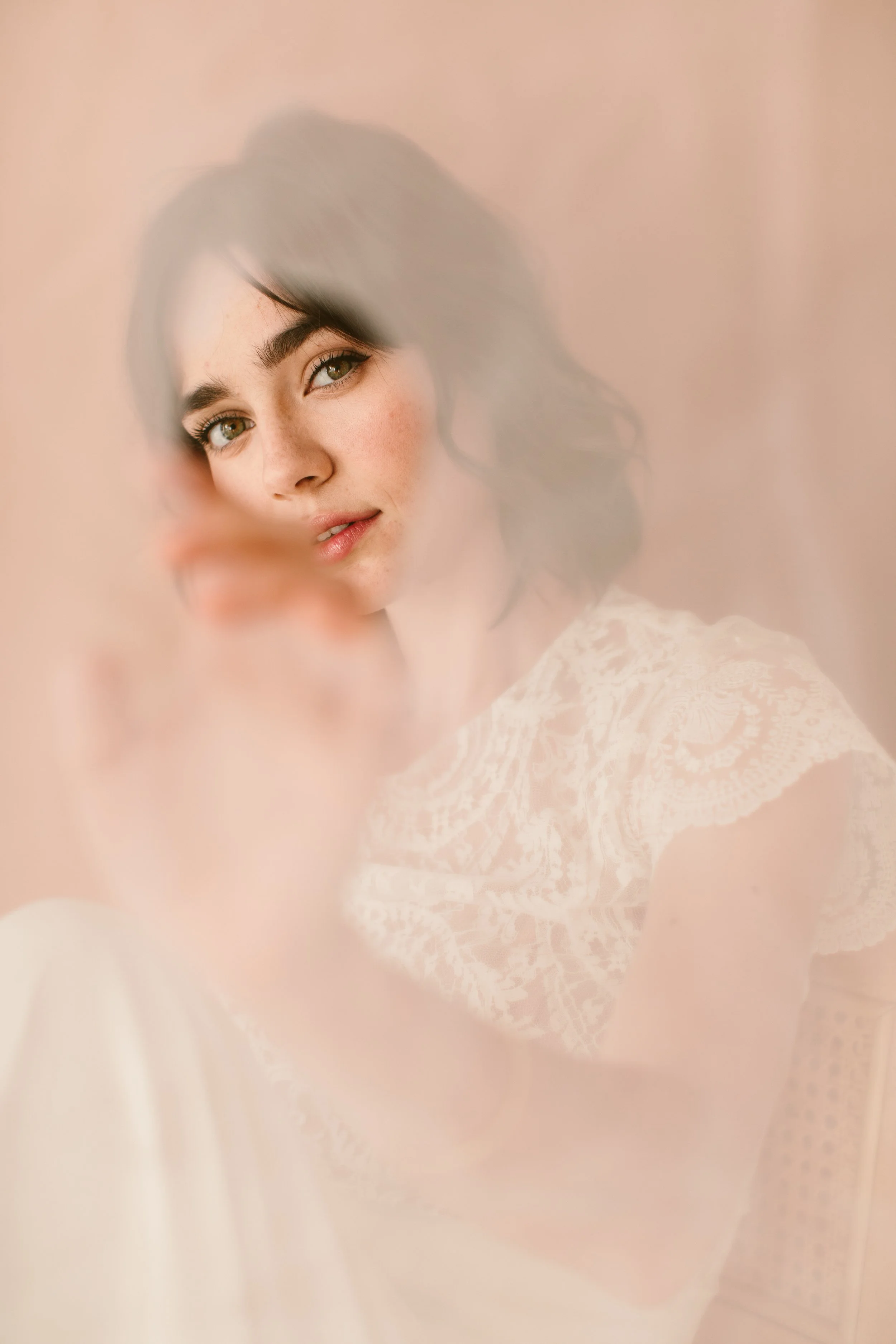 A woman with dark hair and light skin looking through a fuzzy, semi-transparent surface, with a peach-colored background, wearing a lace-patterned outfit.