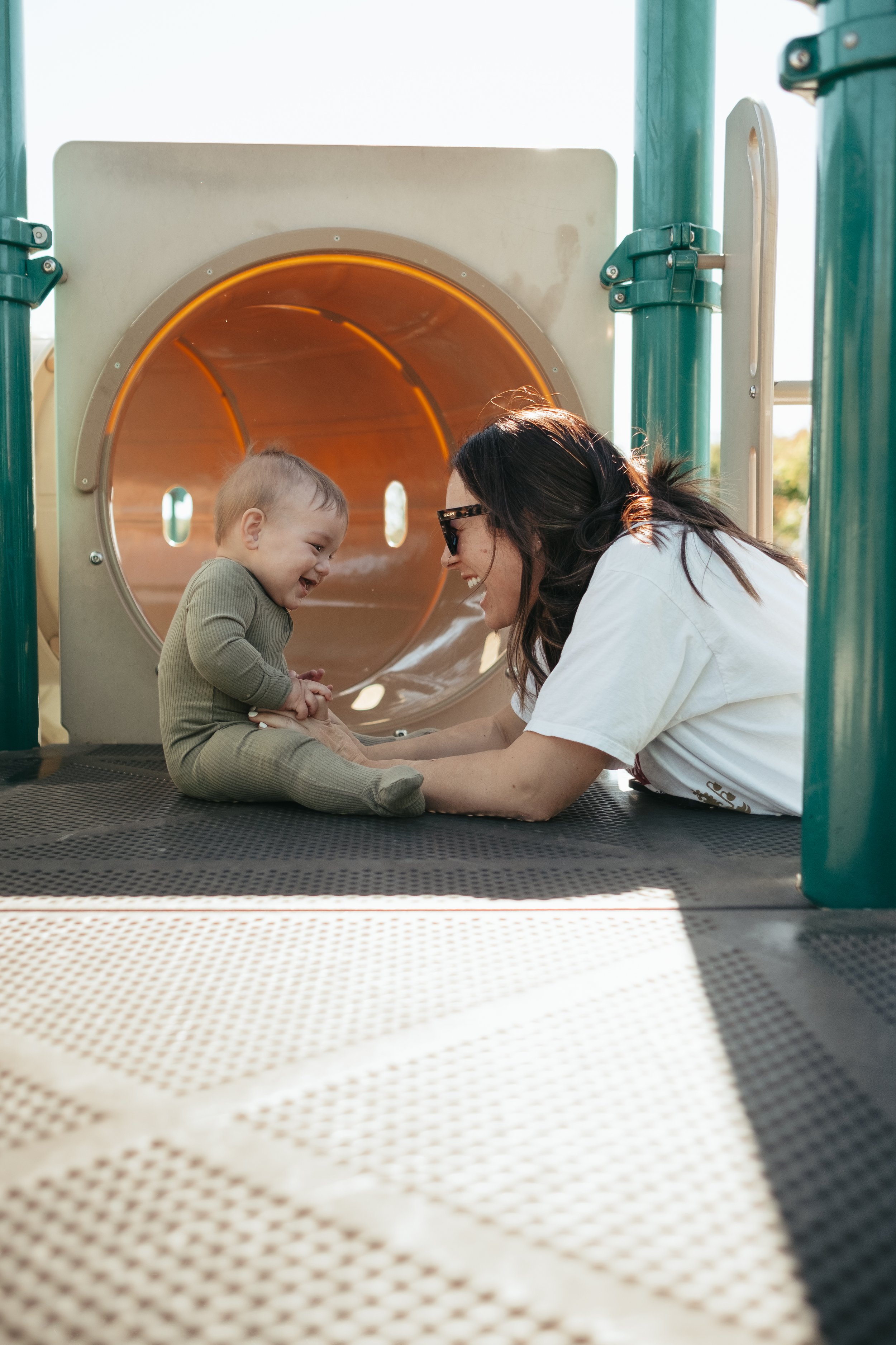 A woman and a young child playing together on a playground structure in Boise, Idaho., smiling and laughing, with the child sitting inside the tunnel and the woman lying on the ground facing him.