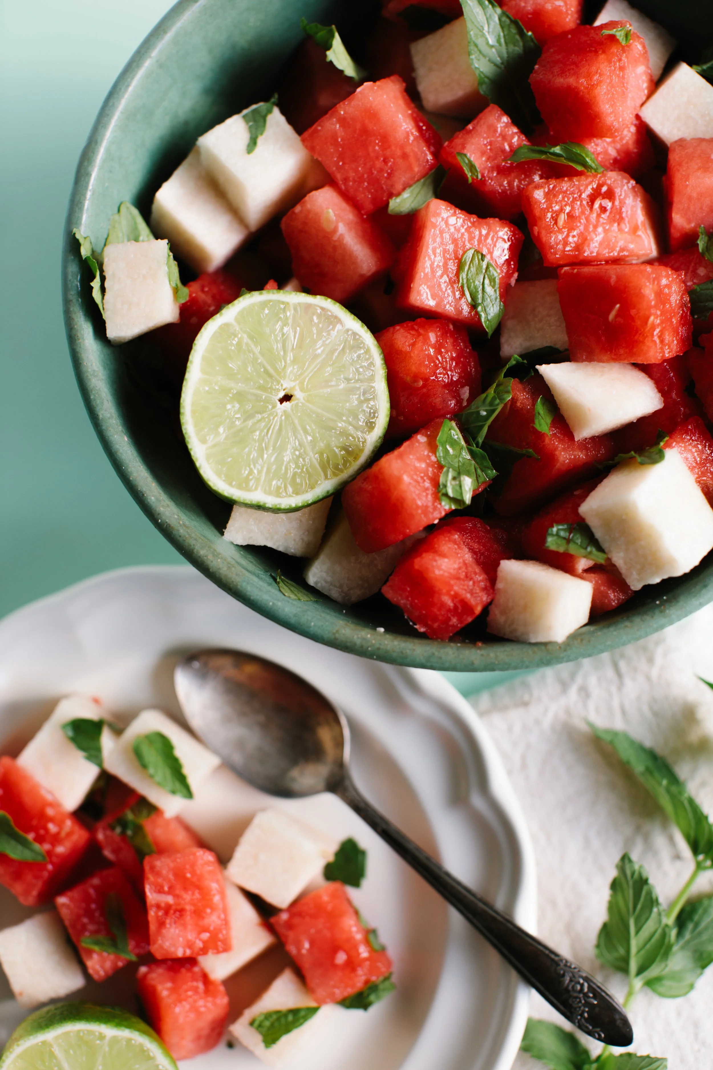 A bowl of watermelon, cantaloupe, and honeydew melon salad garnished with fresh mint, with a slice of lime on top, served with a small plate of the same salad and a vintage spoon.