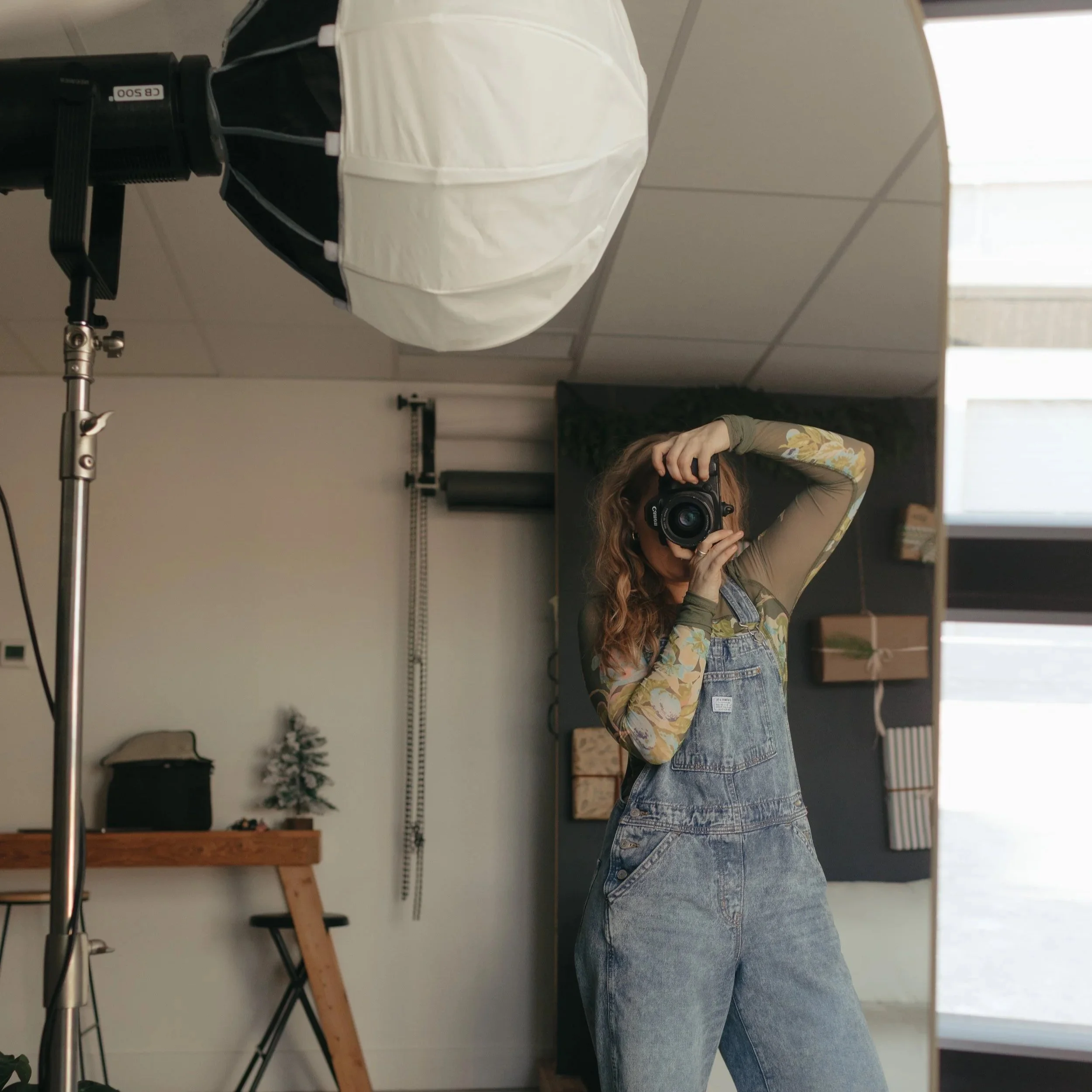 A woman with curly hair, wearing a floral long sleeve shirt and denim overalls, taking a photo with a camera in a photography studio, standing in front of a mirror.