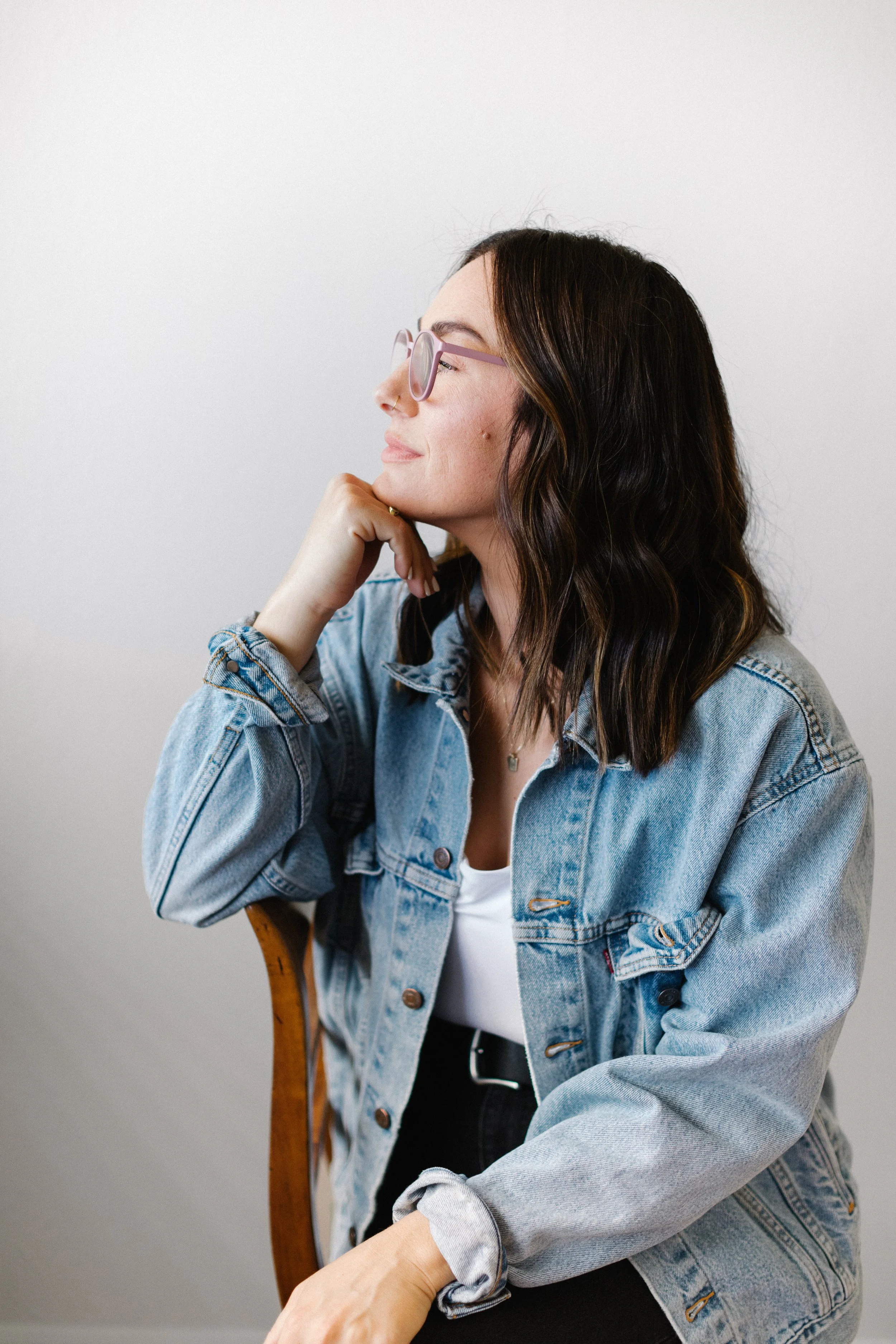 A woman with dark wavy hair and glasses is sitting sideways on a wooden chair with a white background, wearing a blue denim jacket and a white shirt.